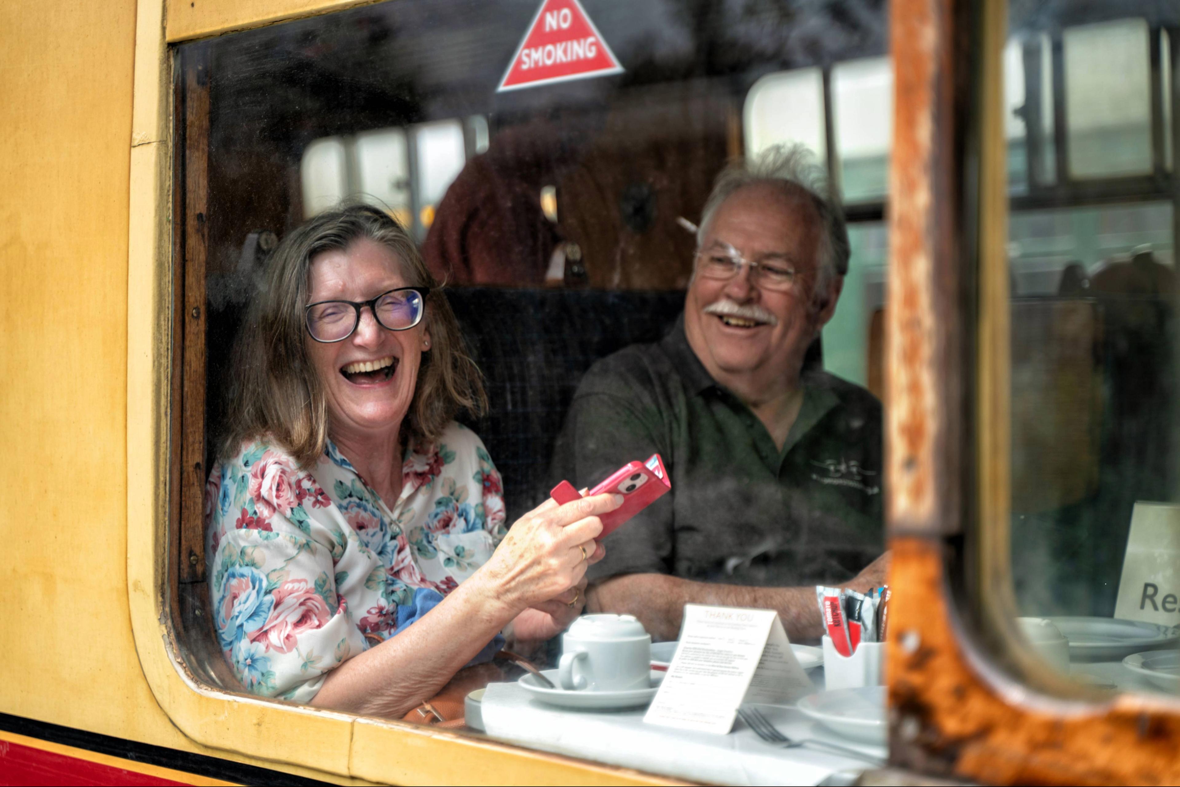 A woman and man laughing together inside a train, surrounded by cups and a “No Smoking” sign visible on the window.