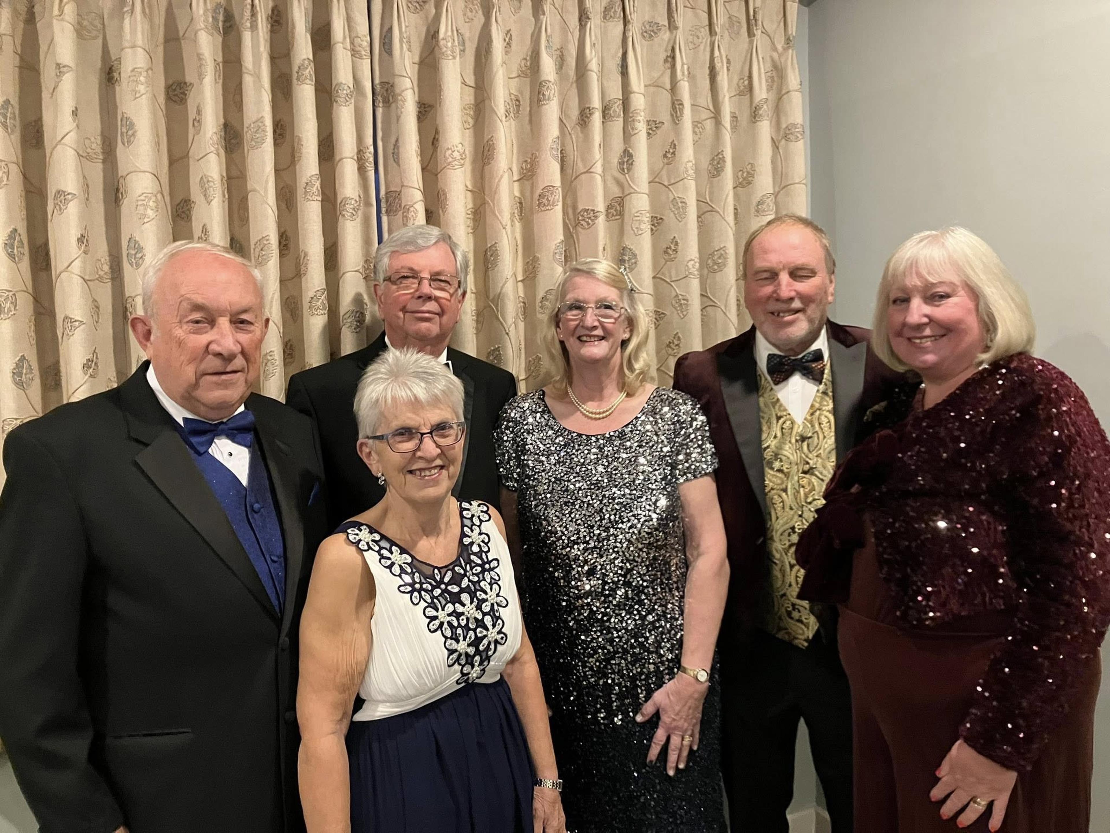 A group of six adults dressed in formal attire, standing in front of patterned curtains