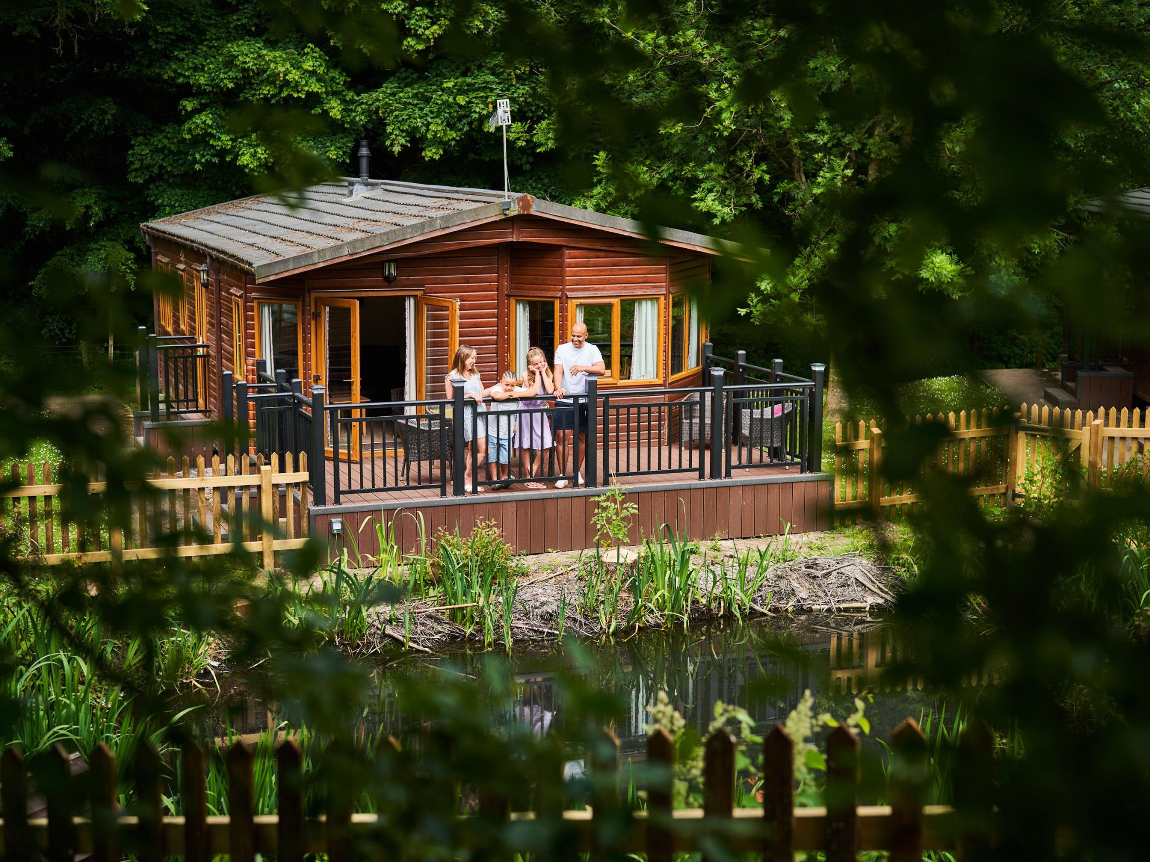 Family on a wooden cabin terrace overlooking a small pond, surrounded by lush greenery and fenced yard.