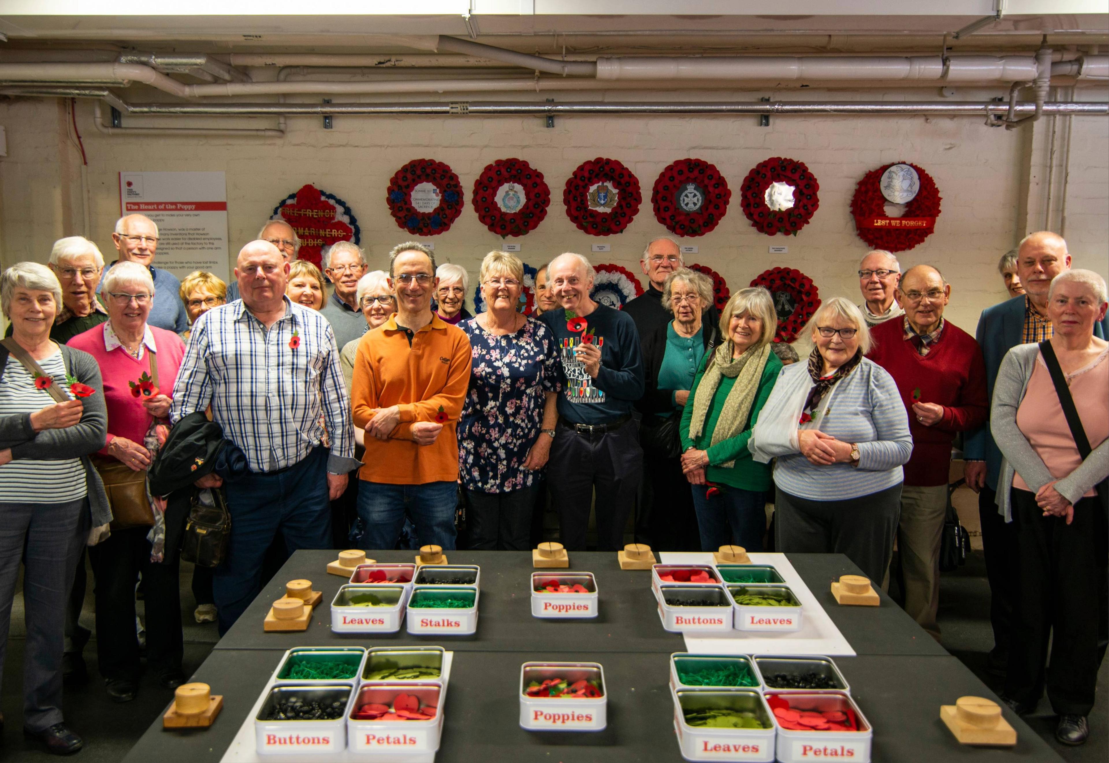 A group of people stands in front of a table with labeled bins of poppy-making materials, with poppy wreaths displayed on the wall behind.
