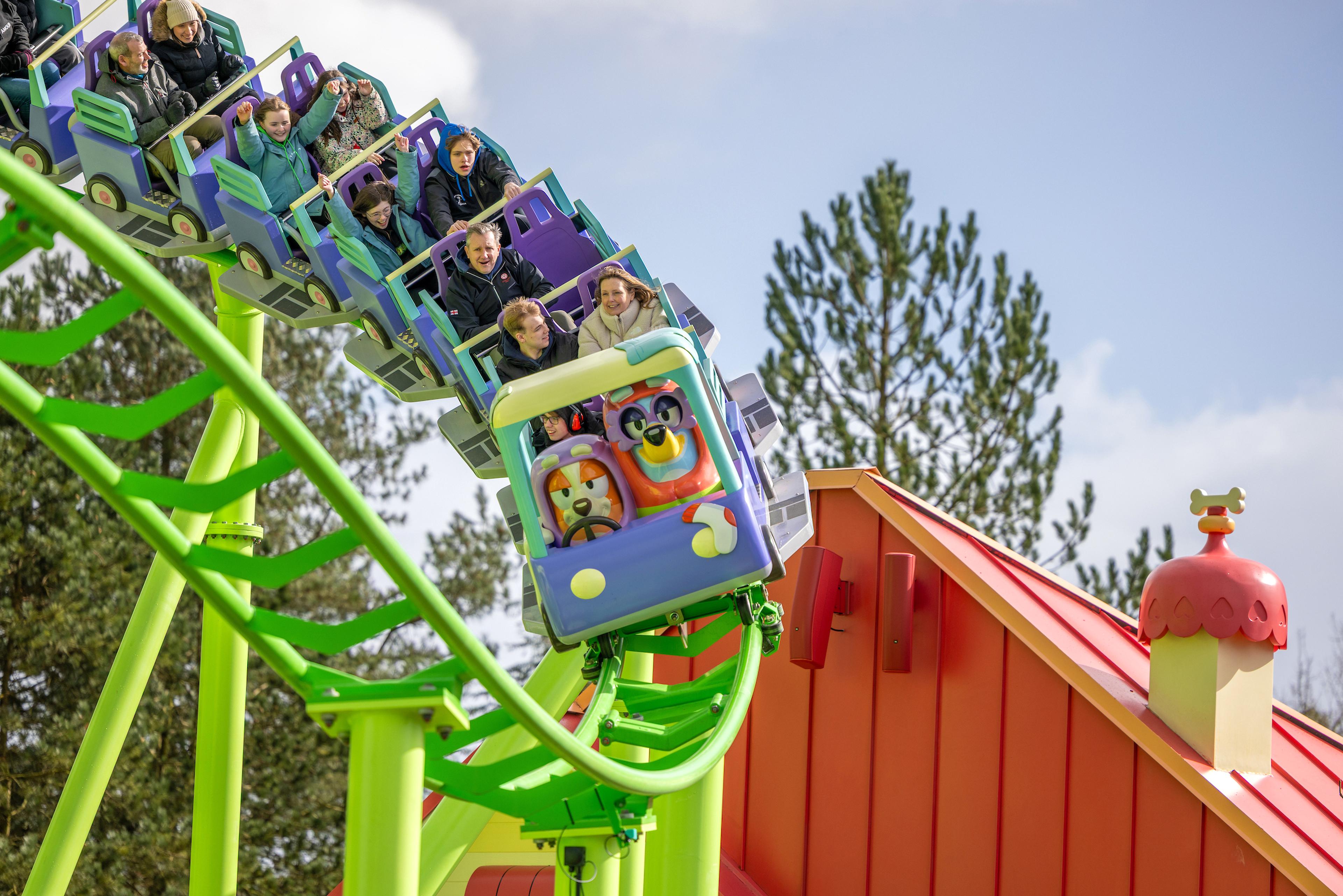 People enjoying a vibrant, green roller coaster ride with colorful train carriages and a playful house structure in the background.