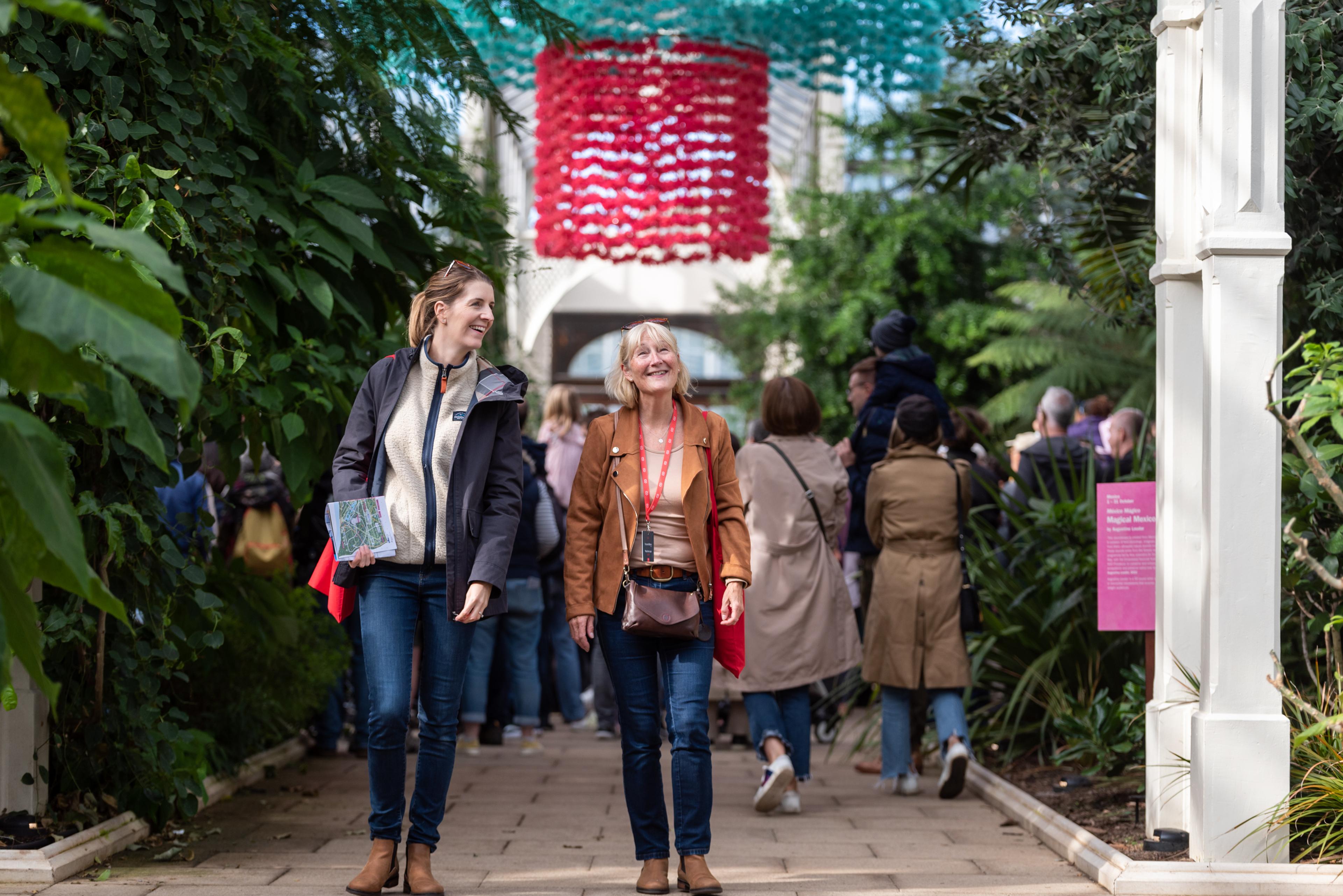 Two women walk through a greenhouse filled with lush greenery, under colorful hanging decorations, amid a group of people.