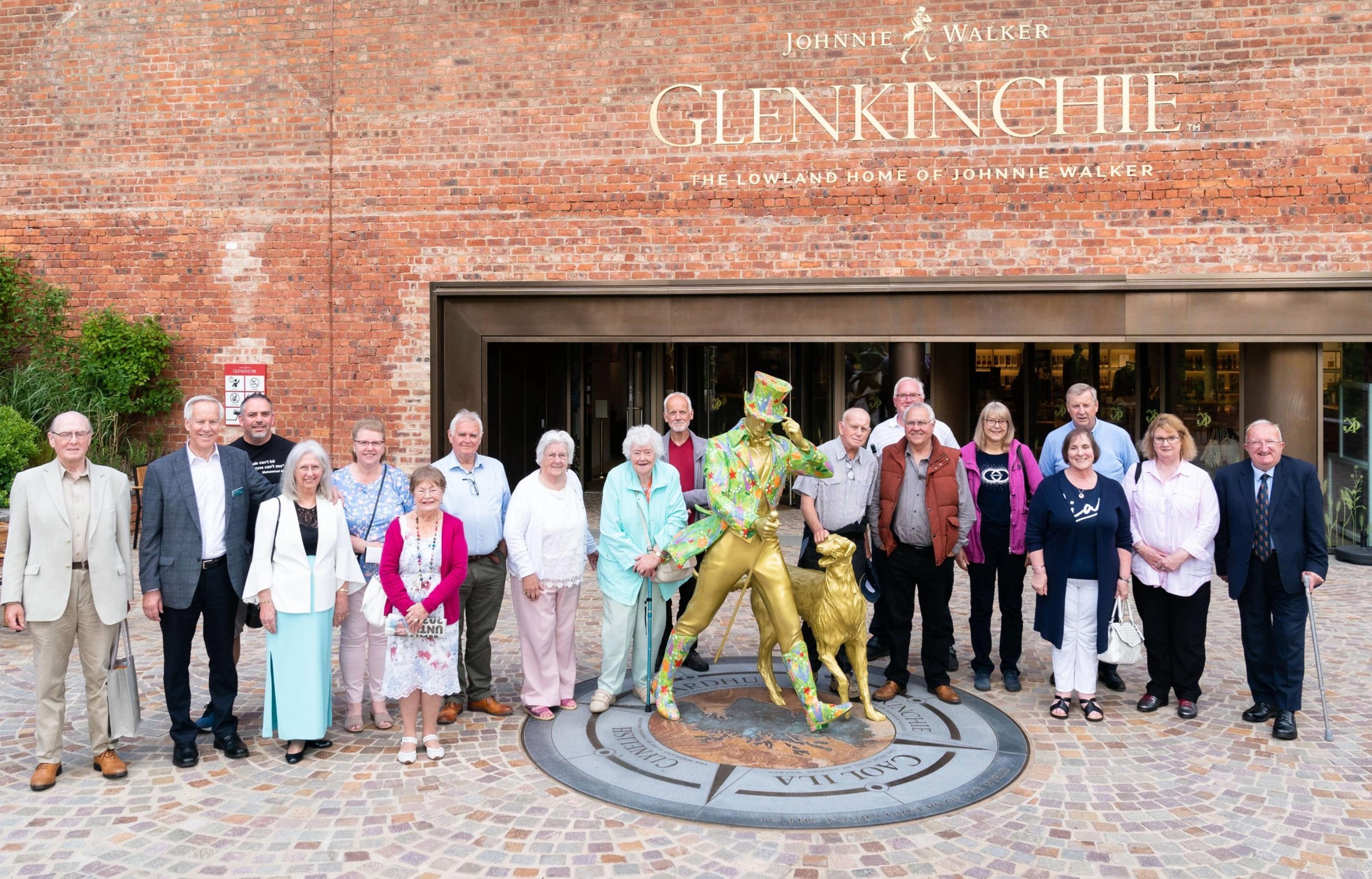 Group of people pose with a colorful statue of Johnnie Walker outside Glenkinchie distillery, with brick building in the background.