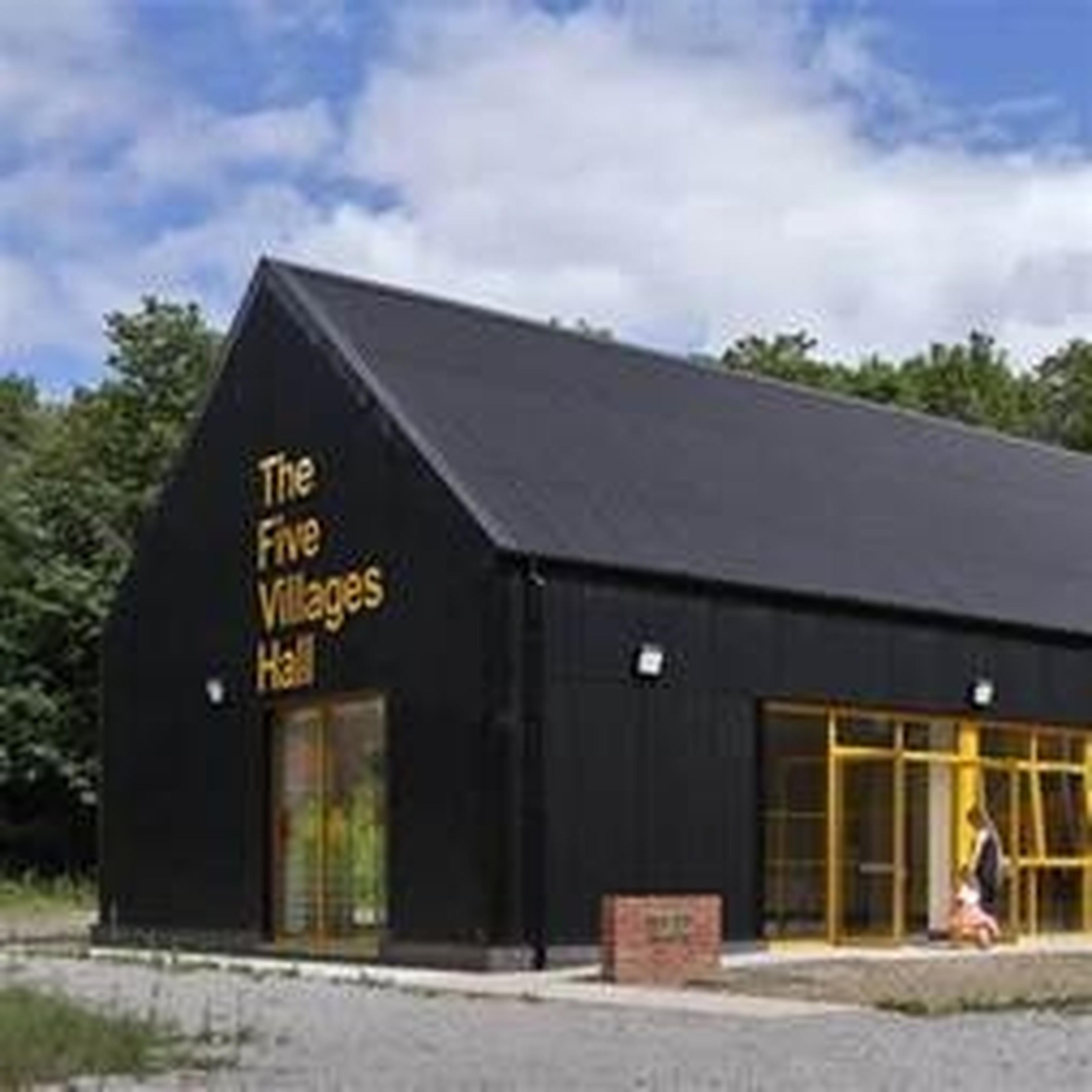 A modern black building labeled "The Five Villages Hall" with yellow doors, set against a backdrop of trees and a cloudy sky.