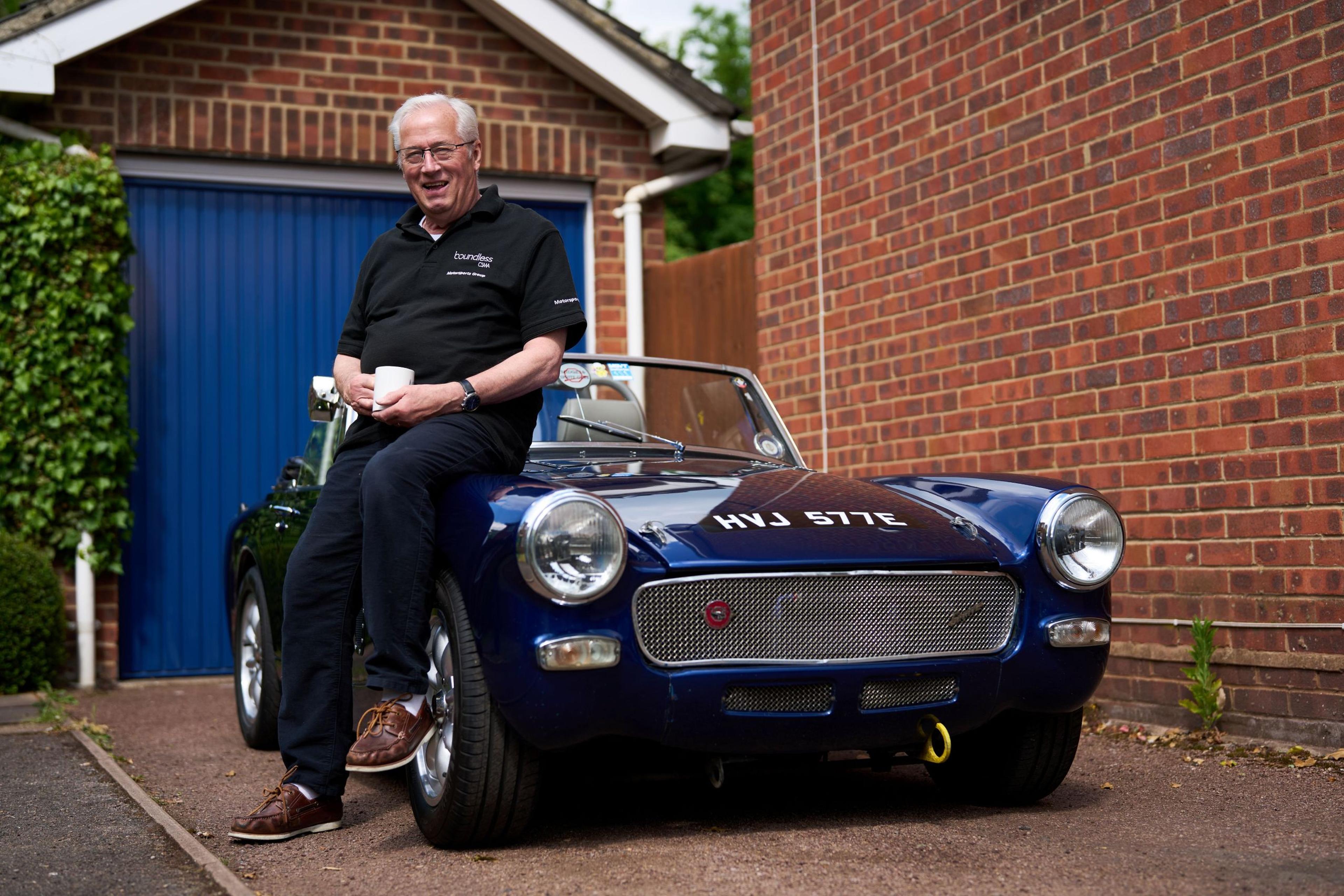 Man sitting on a classic blue convertible, holding a coffee cup, in a driveway with a blue garage door in the background.