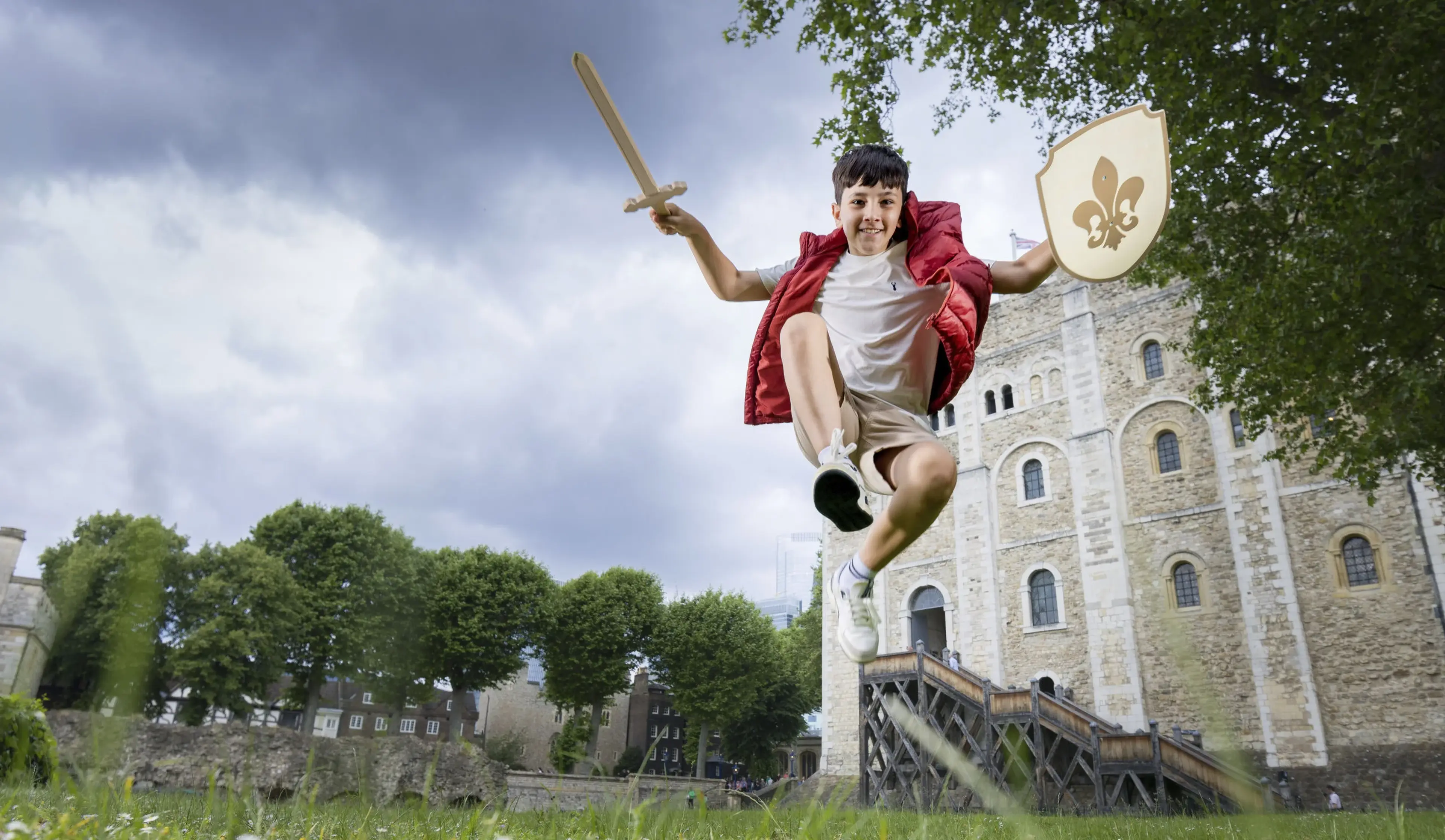 Child in a red vest jumps heroically with a toy sword and shield near a historic stone building, surrounded by greenery and cloudy sky.