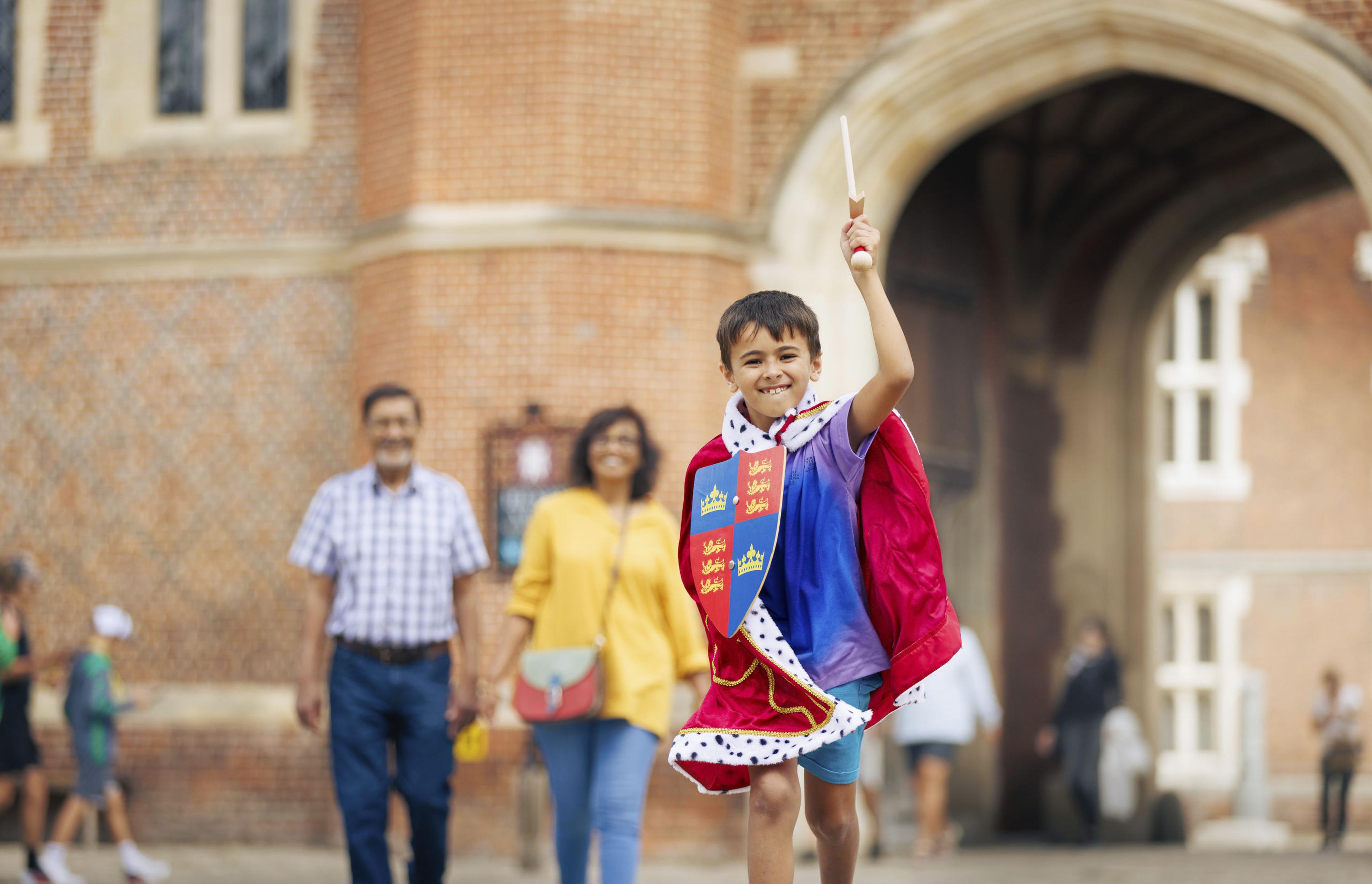 A child in a royal-themed costume joyfully waves a wand. Two adults walk in the background in front of a historical brick building.