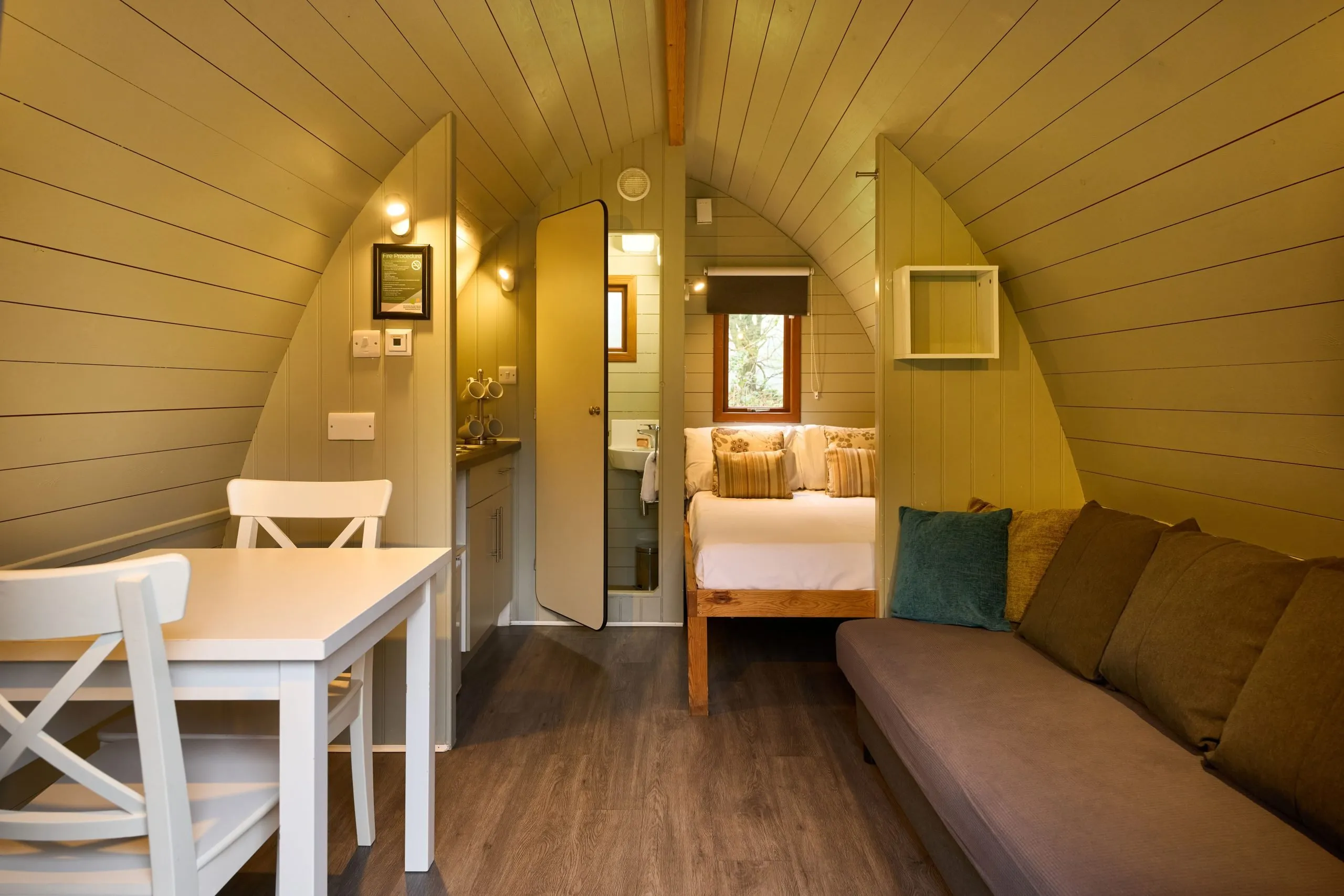 Cozy cabin interior with arched ceiling, featuring a white dining table, a sofa, and a neatly made bed with pillows, all in a warm, wood-paneled space.