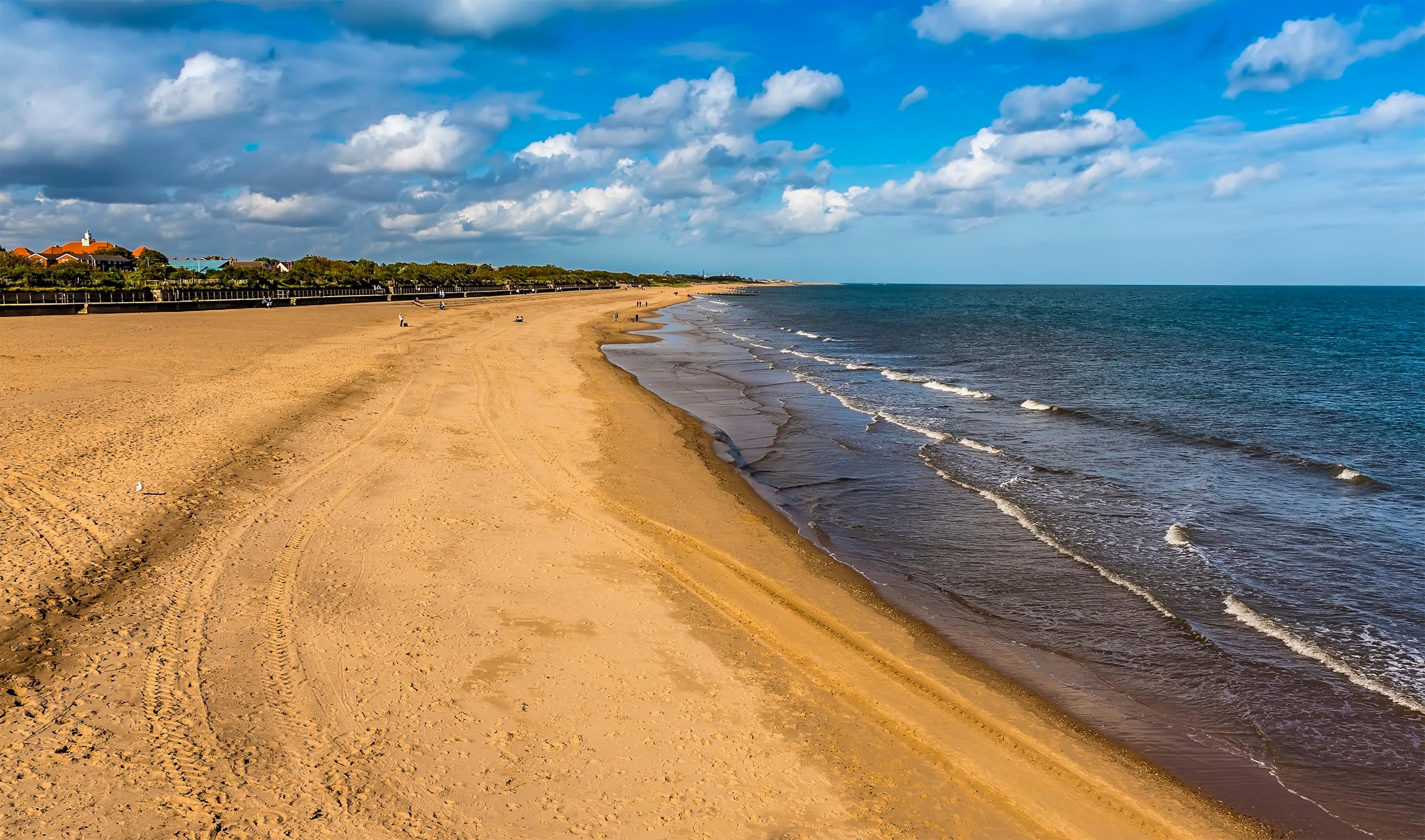 Sandy beach with gentle waves, stretching into the distance under a partly cloudy blue sky, with scattered people in the background.