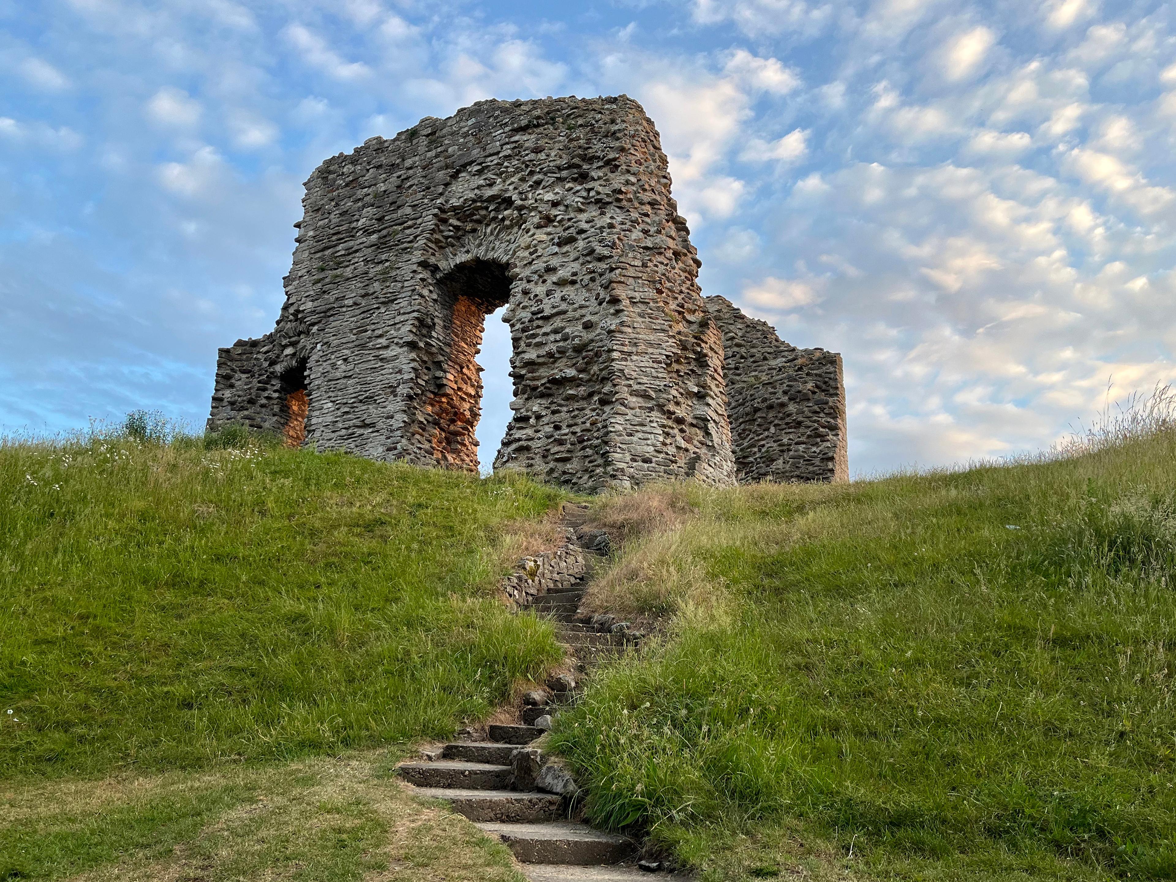 Stone ruins on a grassy hill under a cloudy blue sky, with a stone path leading up to them.