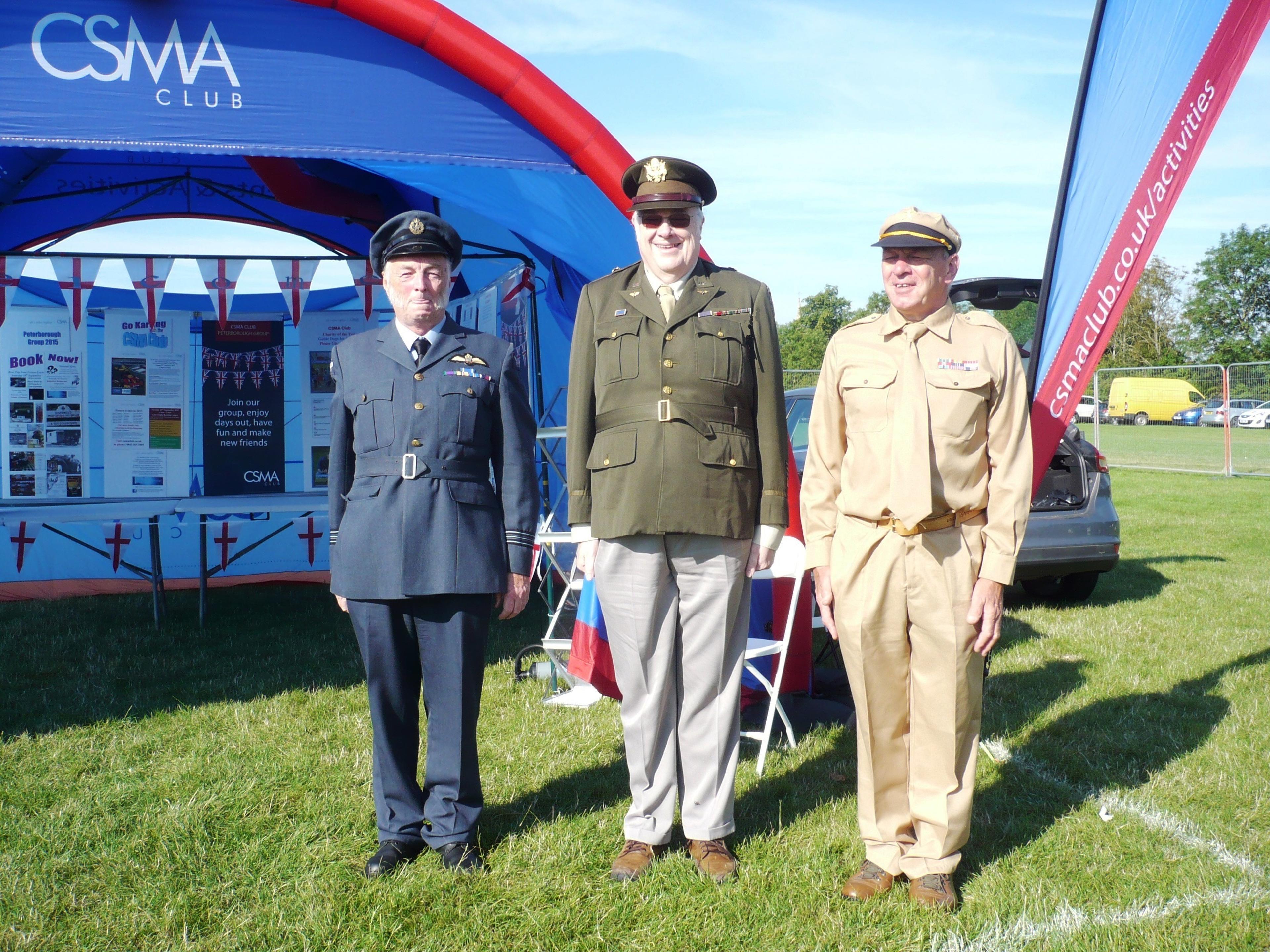 Three men in vintage military uniforms stand in front of a CSMA Club tent
