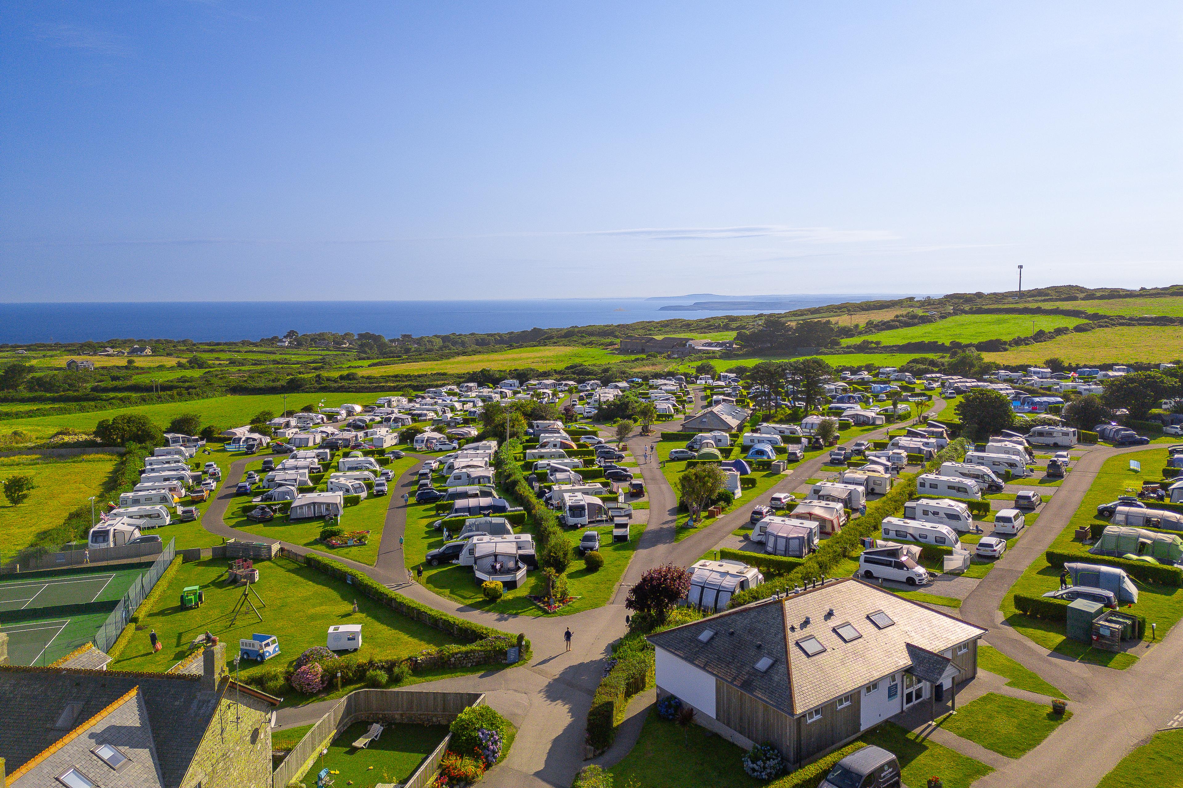 Aerial view of a campsite with numerous caravans and motorhomes on grassy plots near the coastline, under a clear blue sky.