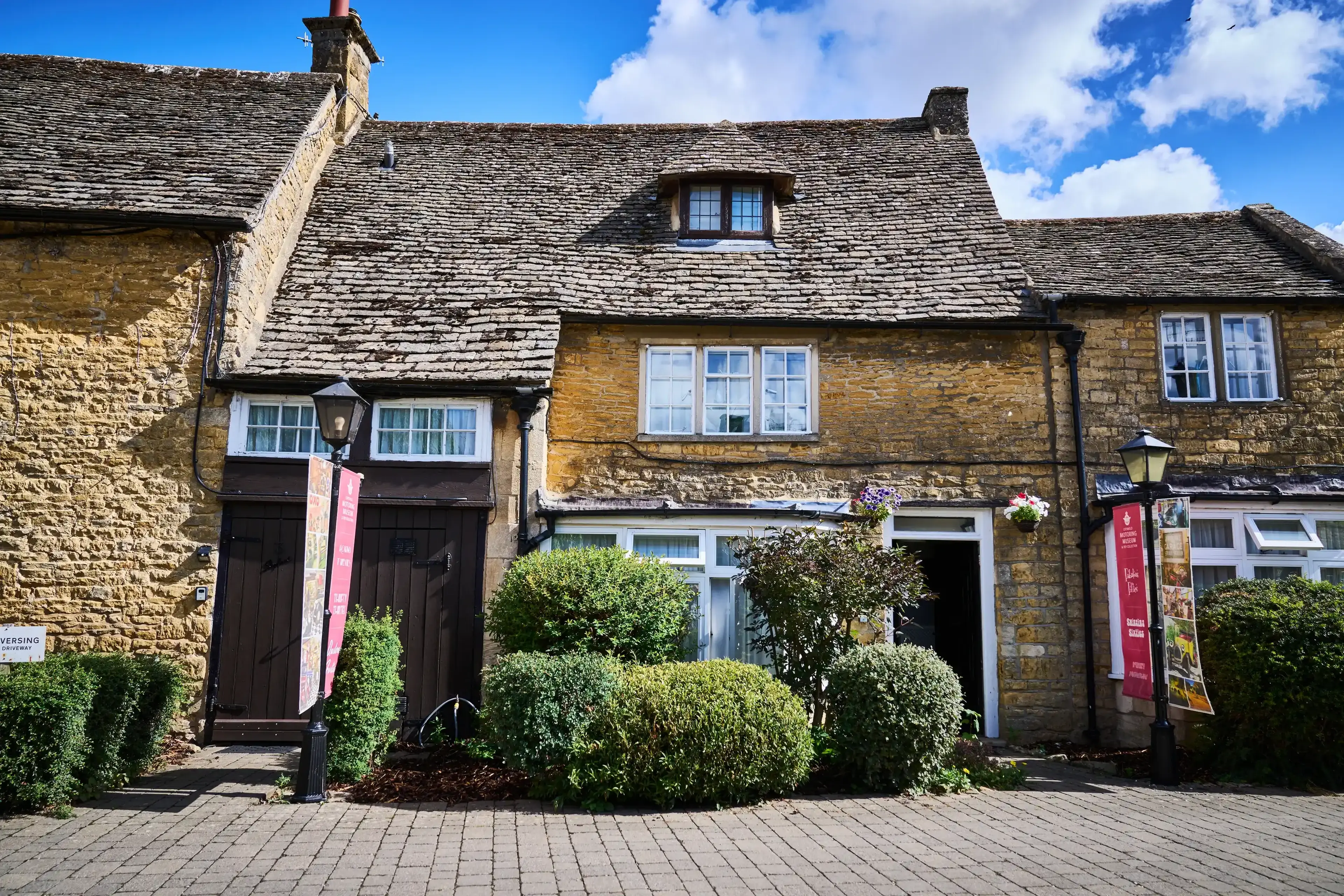 Charming stone cottage with a sloped roof, colorful banners, and lush bushes lining the cobblestone path under a clear blue sky.