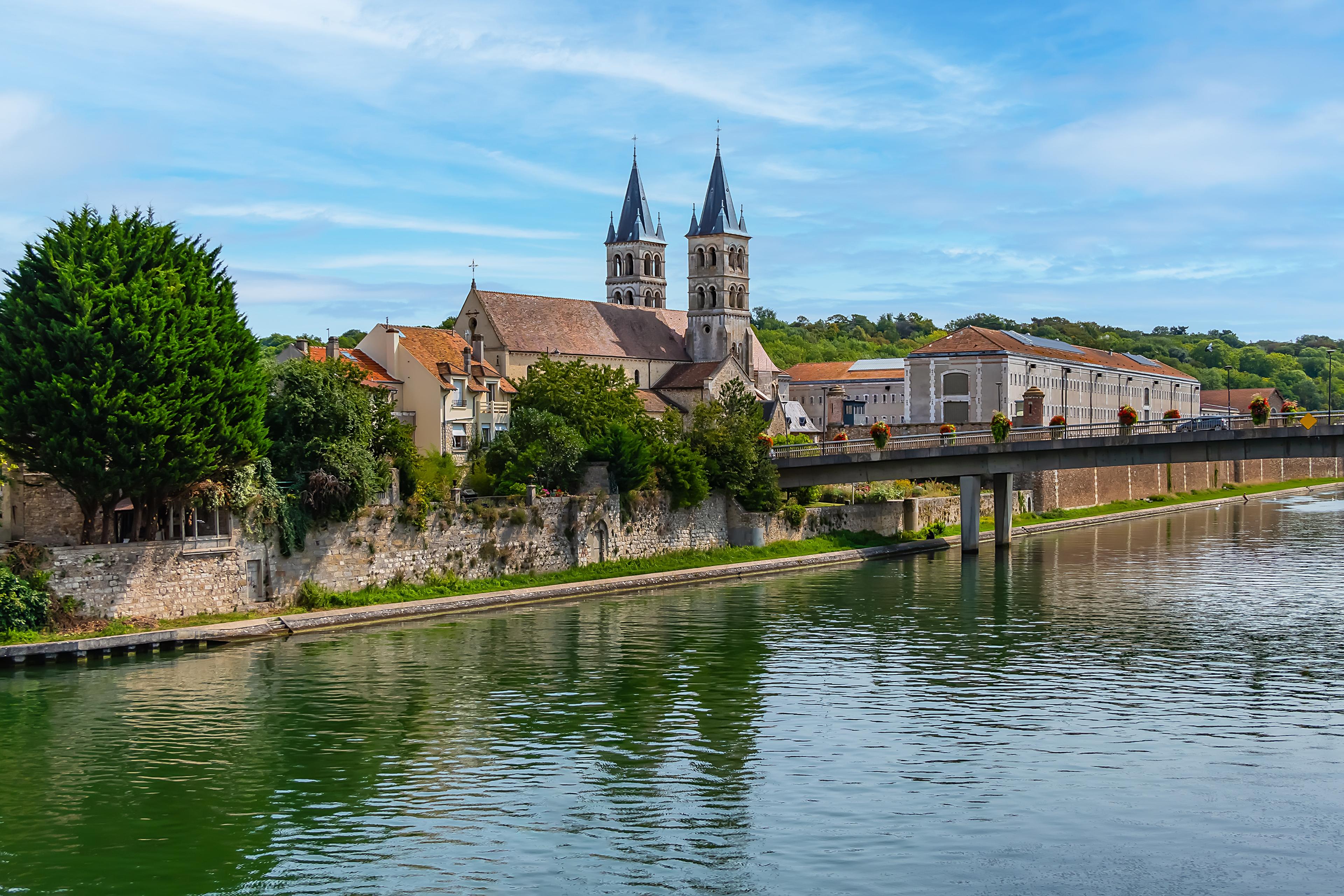 A scenic view of a riverside town with a historic church featuring twin spires, surrounded by trees and old buildings, under a blue sky.