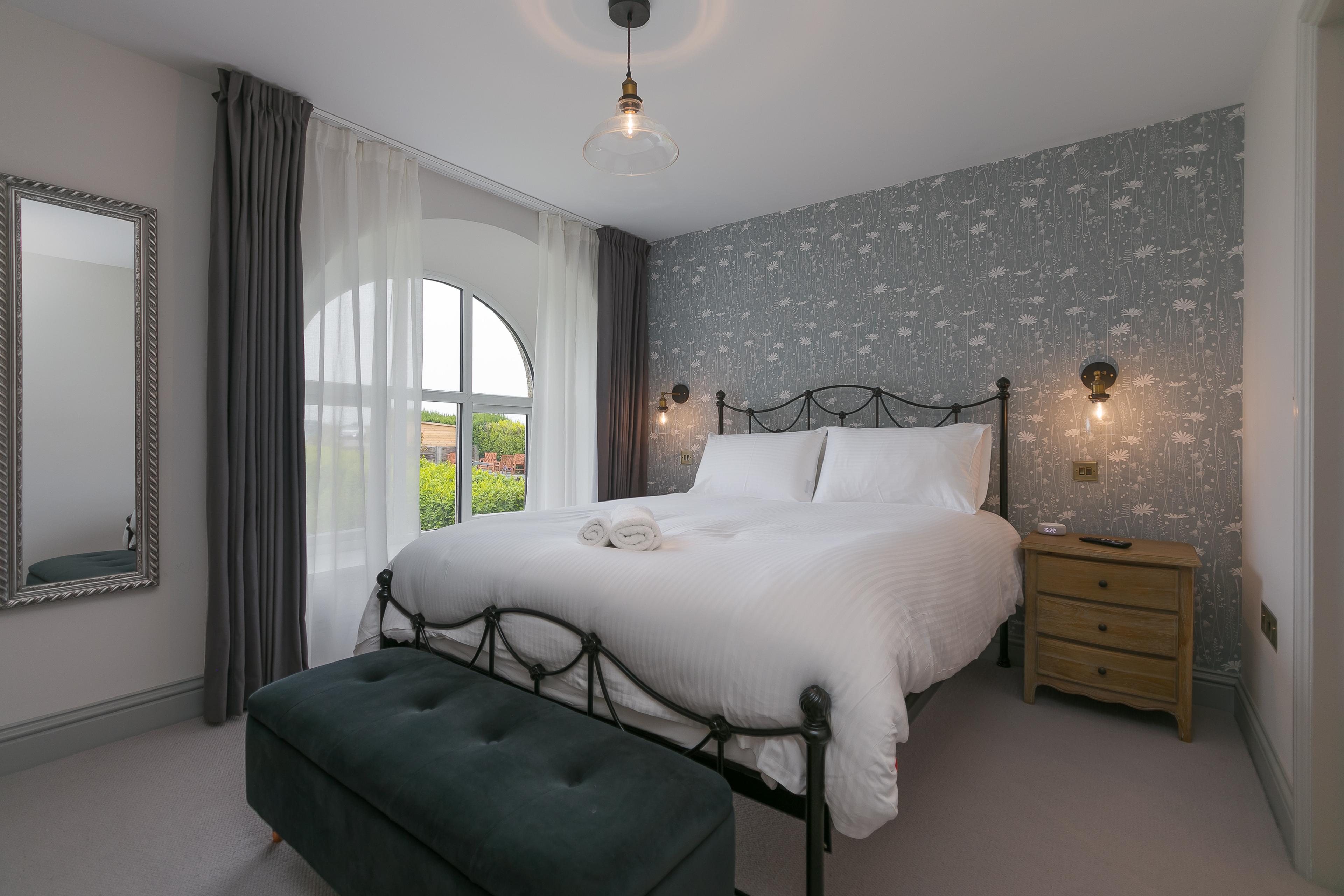 Cozy bedroom with a double bed, floral wallpaper, arched window, and a wooden nightstand. A bench sits at the foot of the bed.