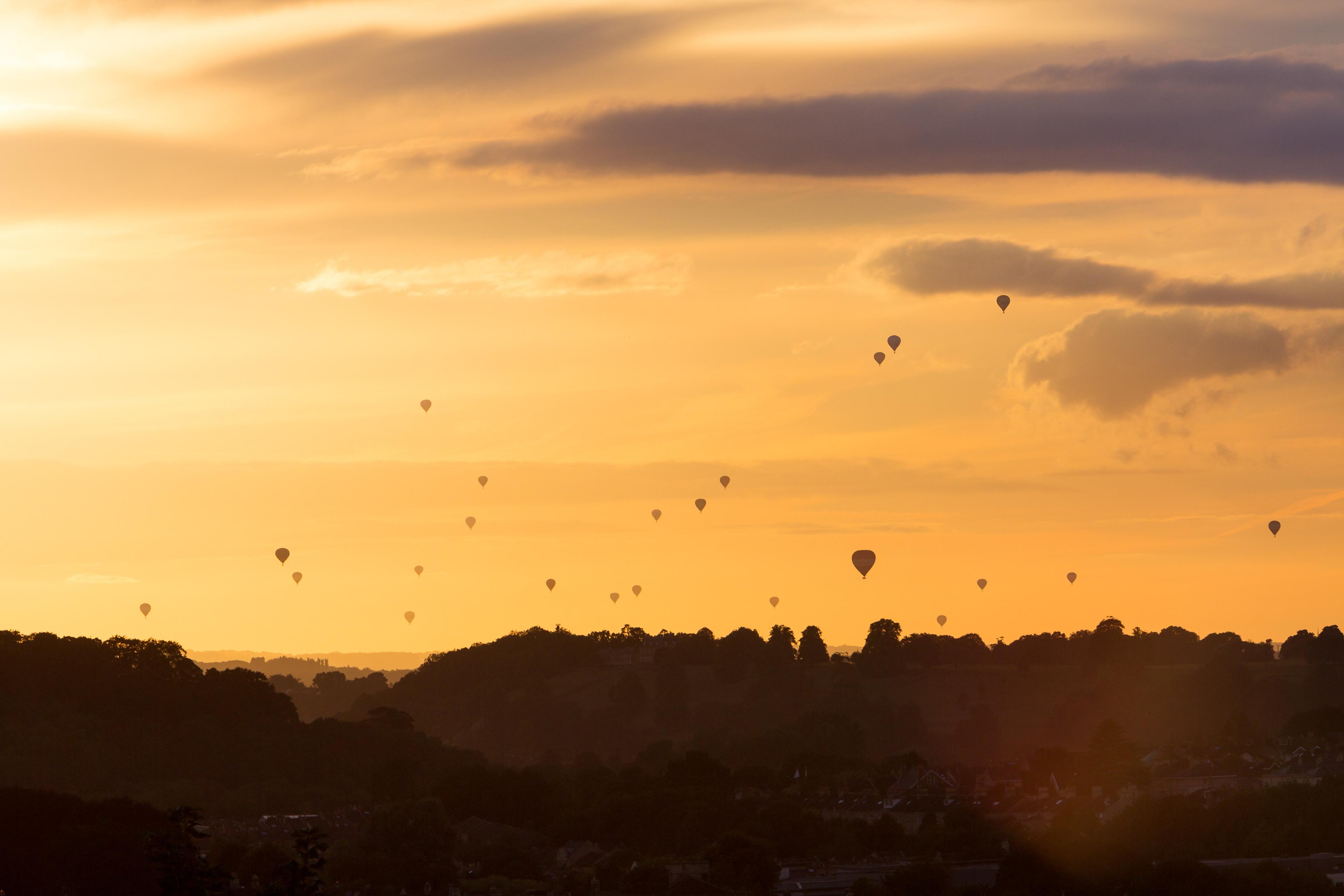 Golden sunset over silhouetted rolling hills, with scattered hot air balloons rising through glowing clouds, creating a peaceful, dreamy atmosphere.