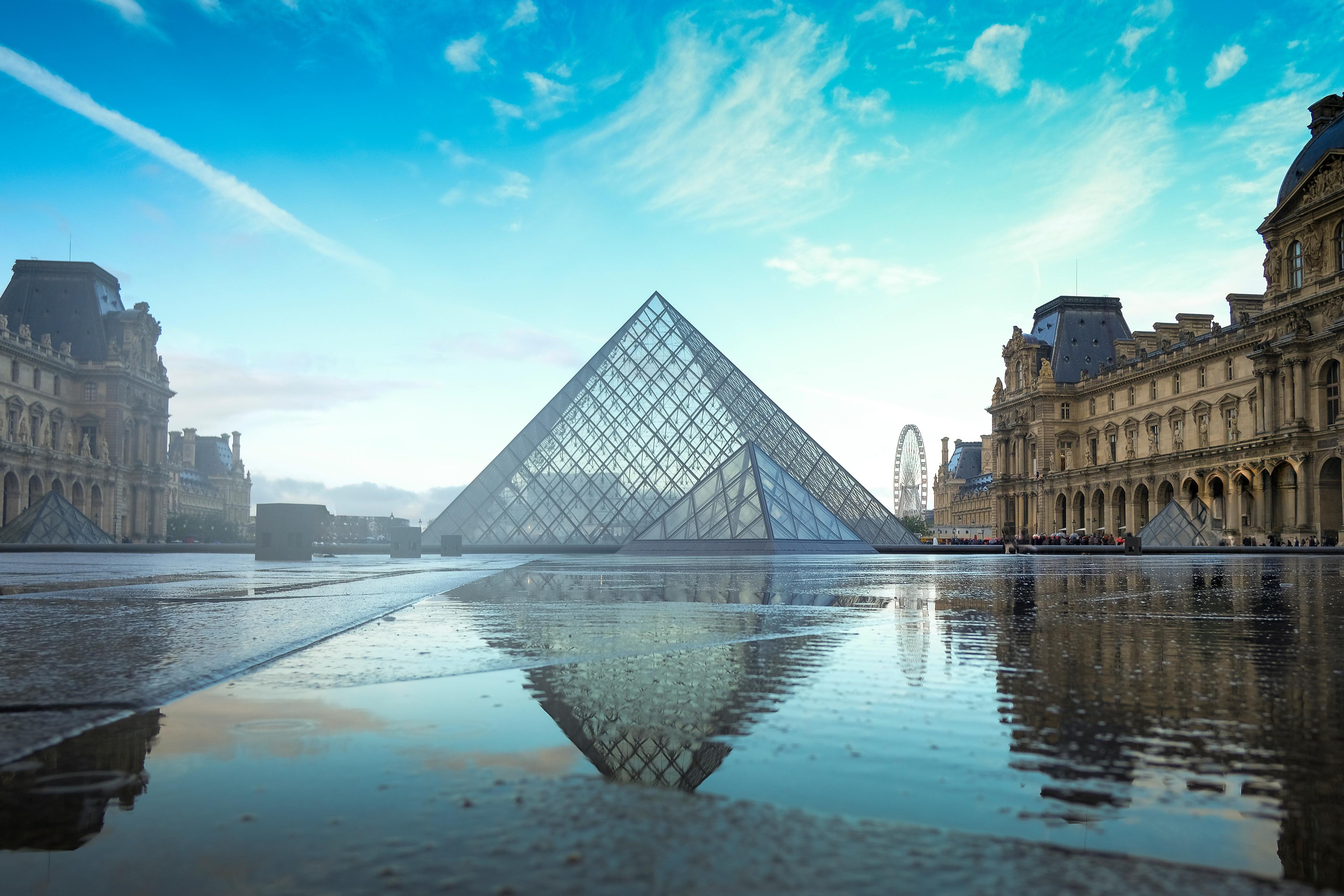 The Louvre Pyramid reflected on wet pavement under a blue sky, surrounded by historic architecture in Paris.