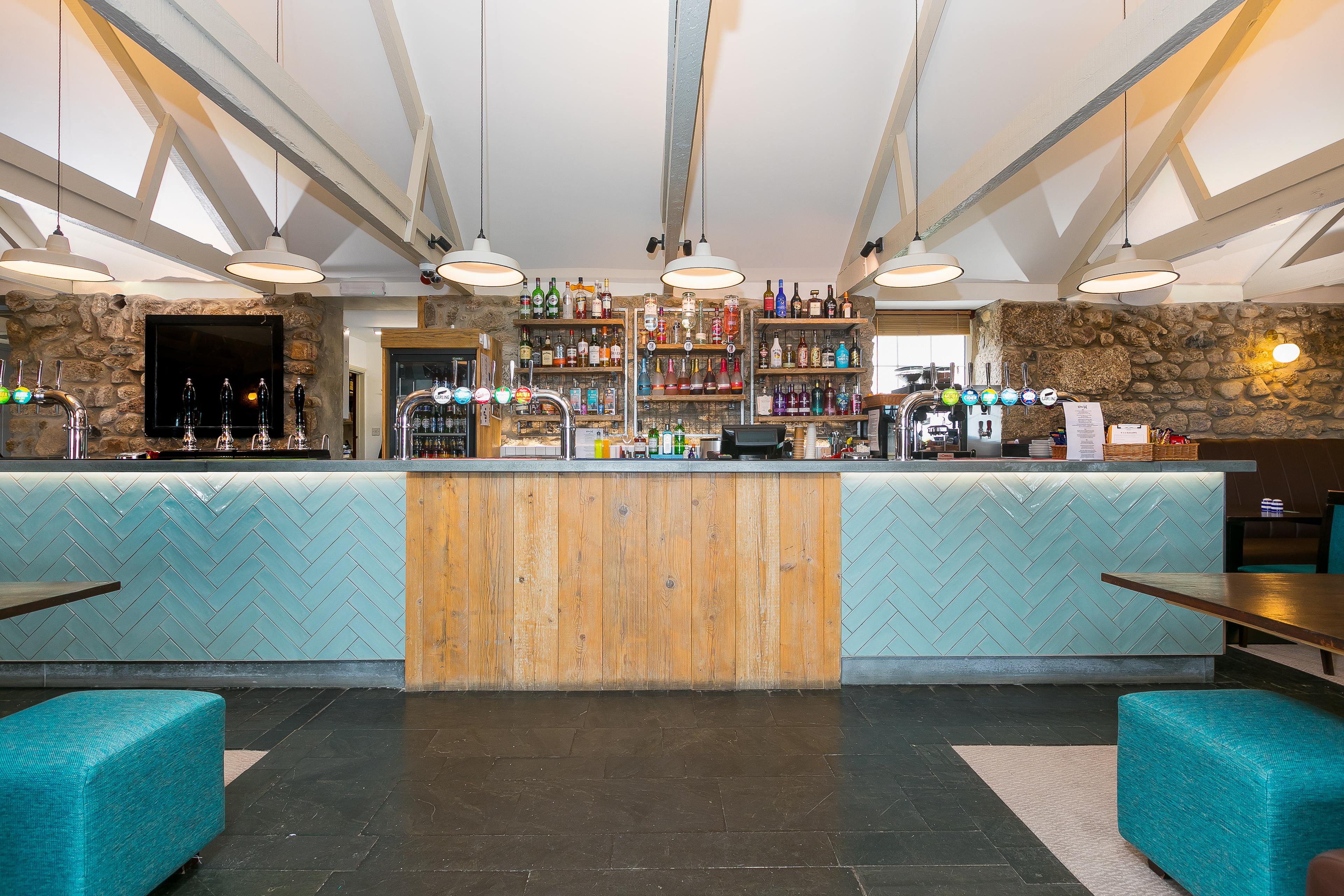 Modern bar interior with a stone wall, wood accents, and blue herringbone tiles. Shelves display various drinks behind the counter.