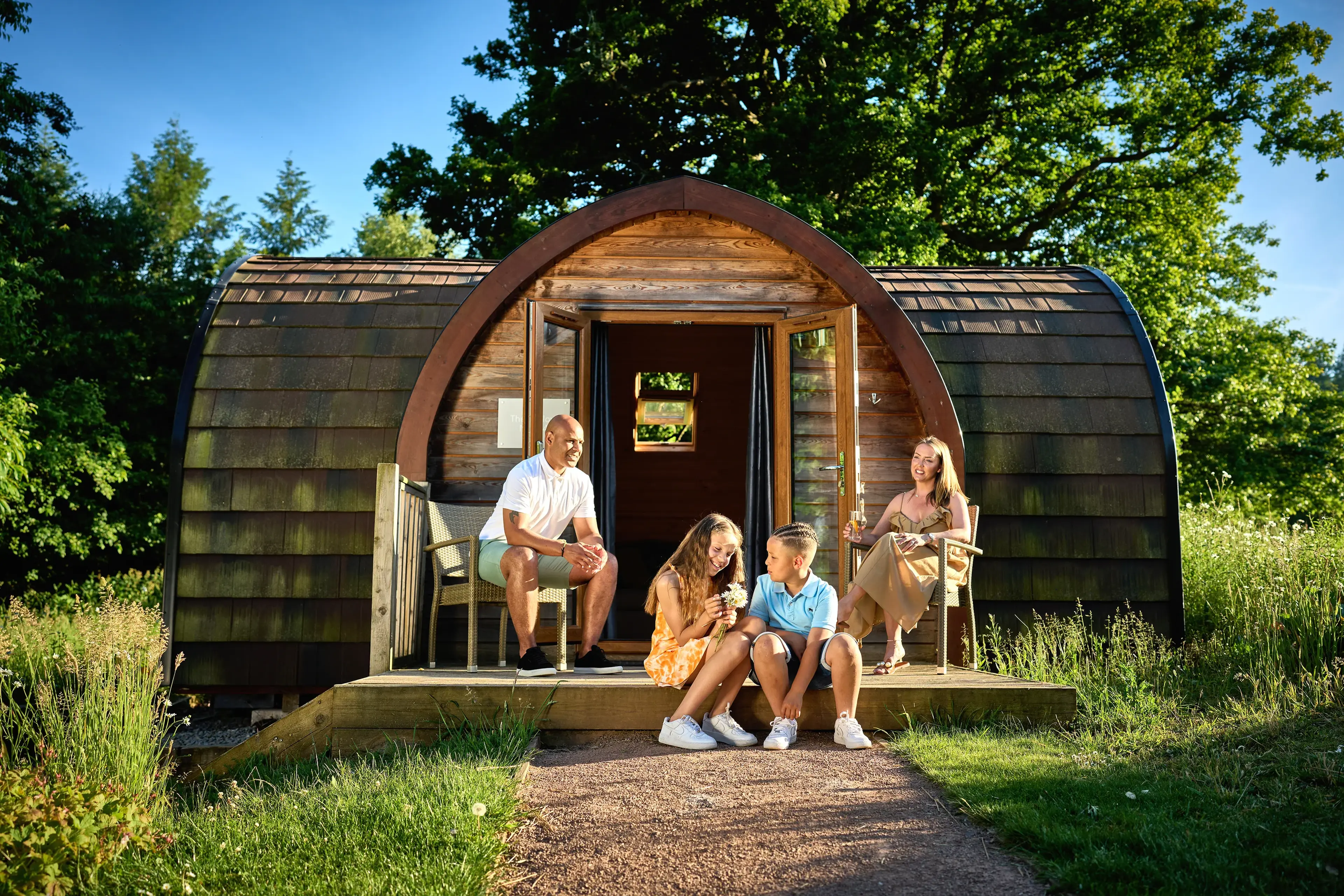 A family of four sits on the porch of a wooden cabin in a lush, green setting under clear skies, enjoying a sunny day together.