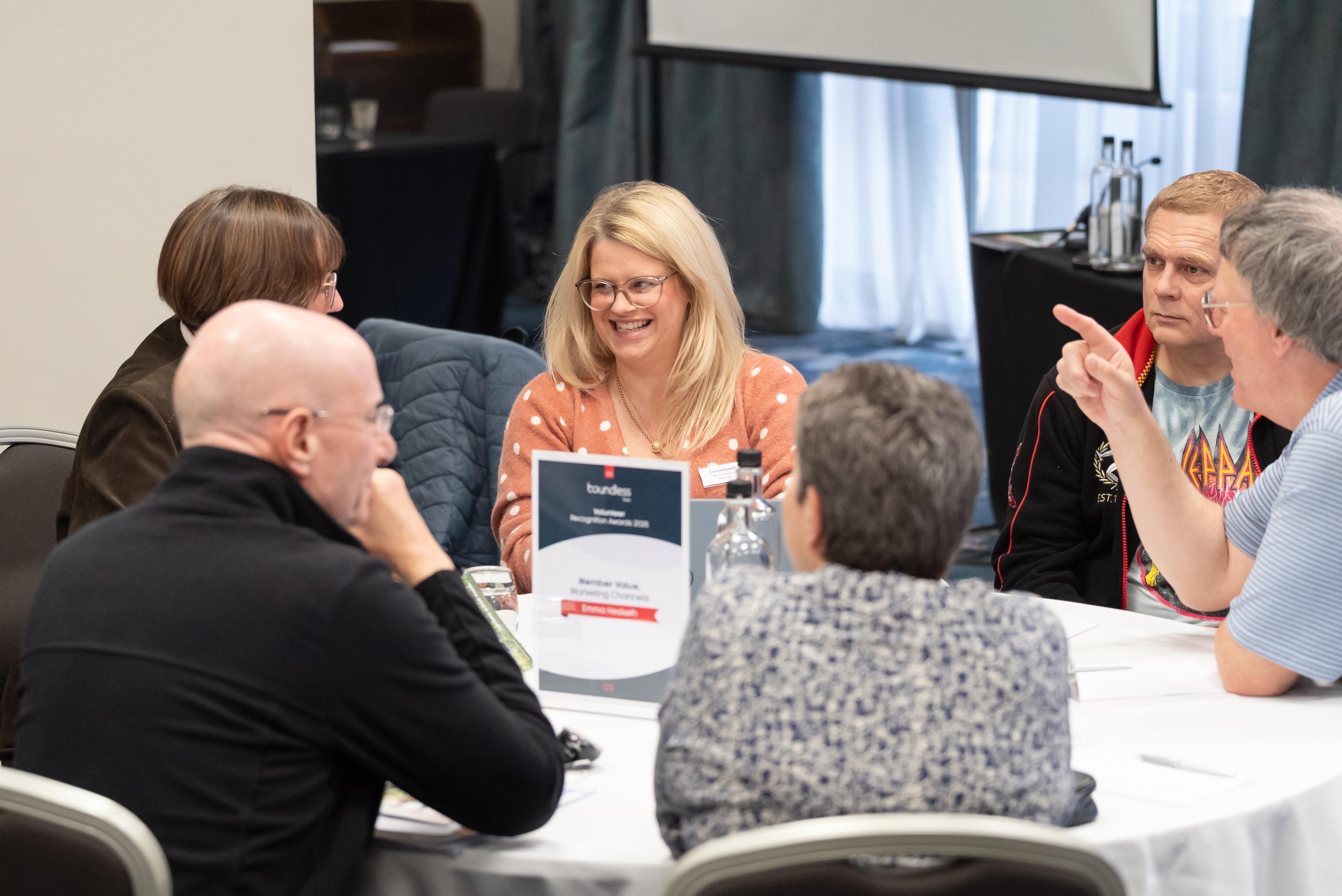 A group of people are sitting around a table in discussion, with a woman in a polka dot sweater smiling. A sign on the table indicates a meeting.