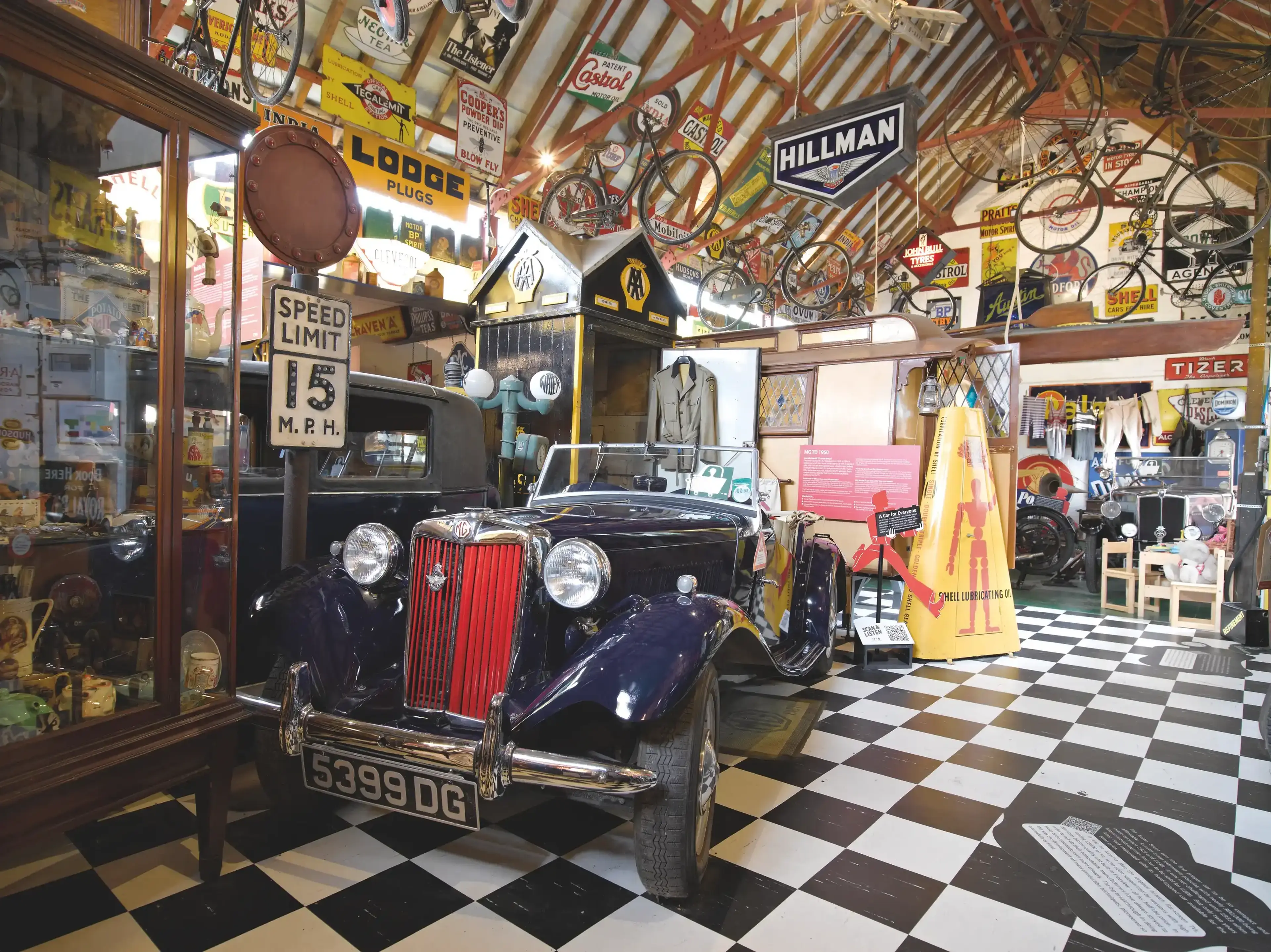 Vintage car museum with a classic black MG convertible, retro signs, bicycles, and memorabilia on display in a checkered floor room.