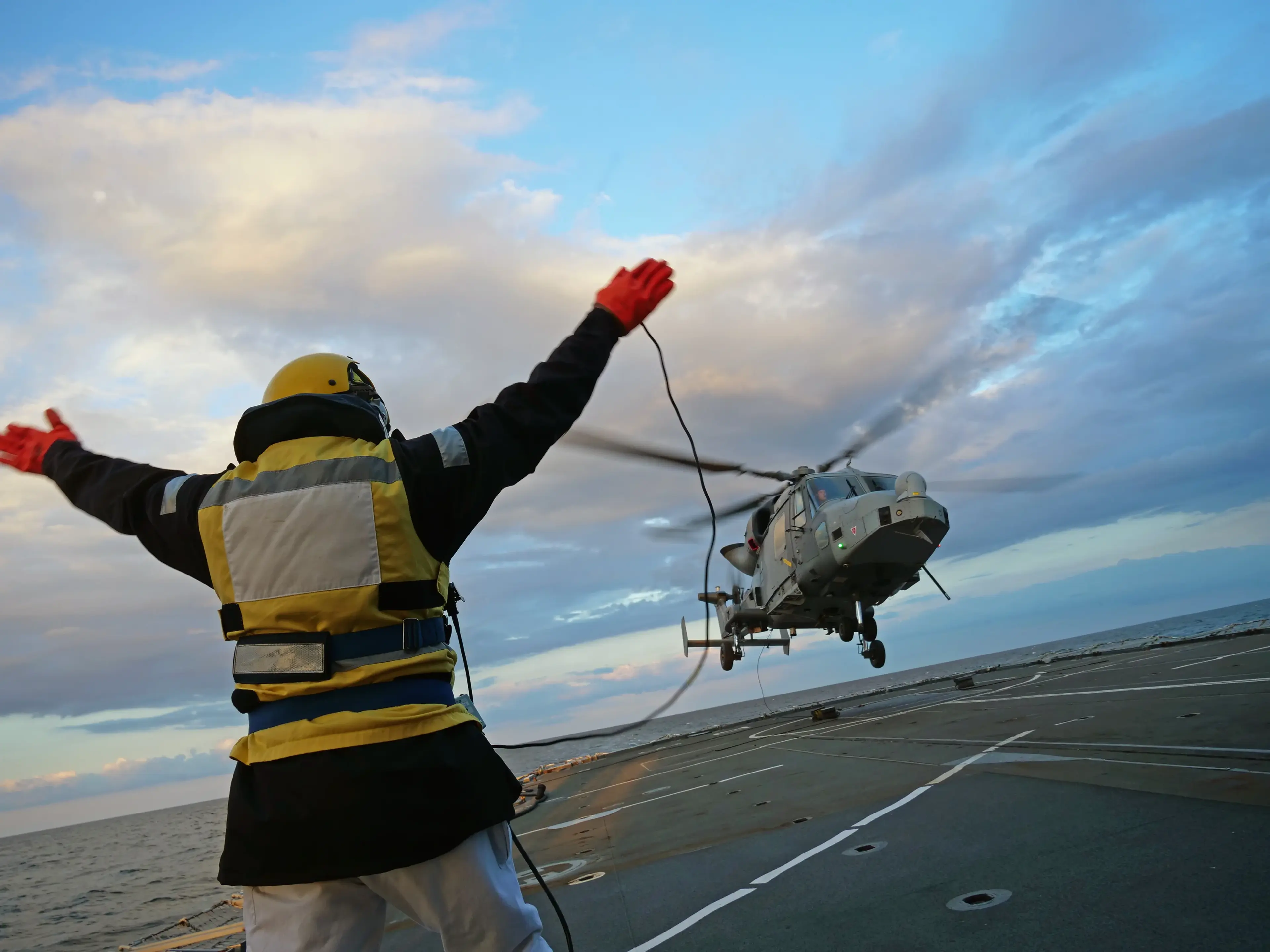 A deck crew member signals a helicopter landing on a ship at sea, with arms outstretched against a cloudy sky backdrop.