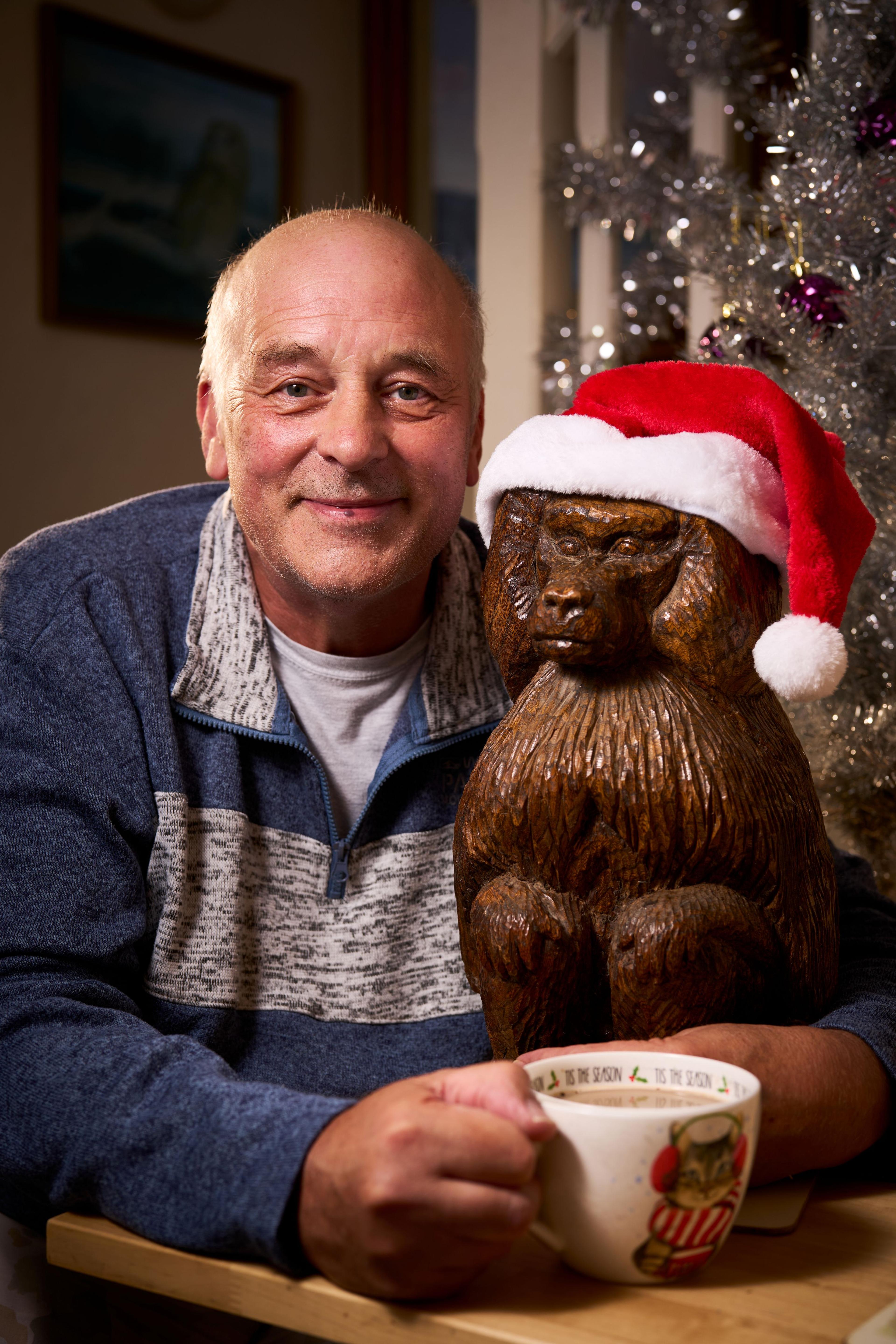 A man holding a cup of tea sitting next to a wooden sculpture of a baboon wearing a santa hat