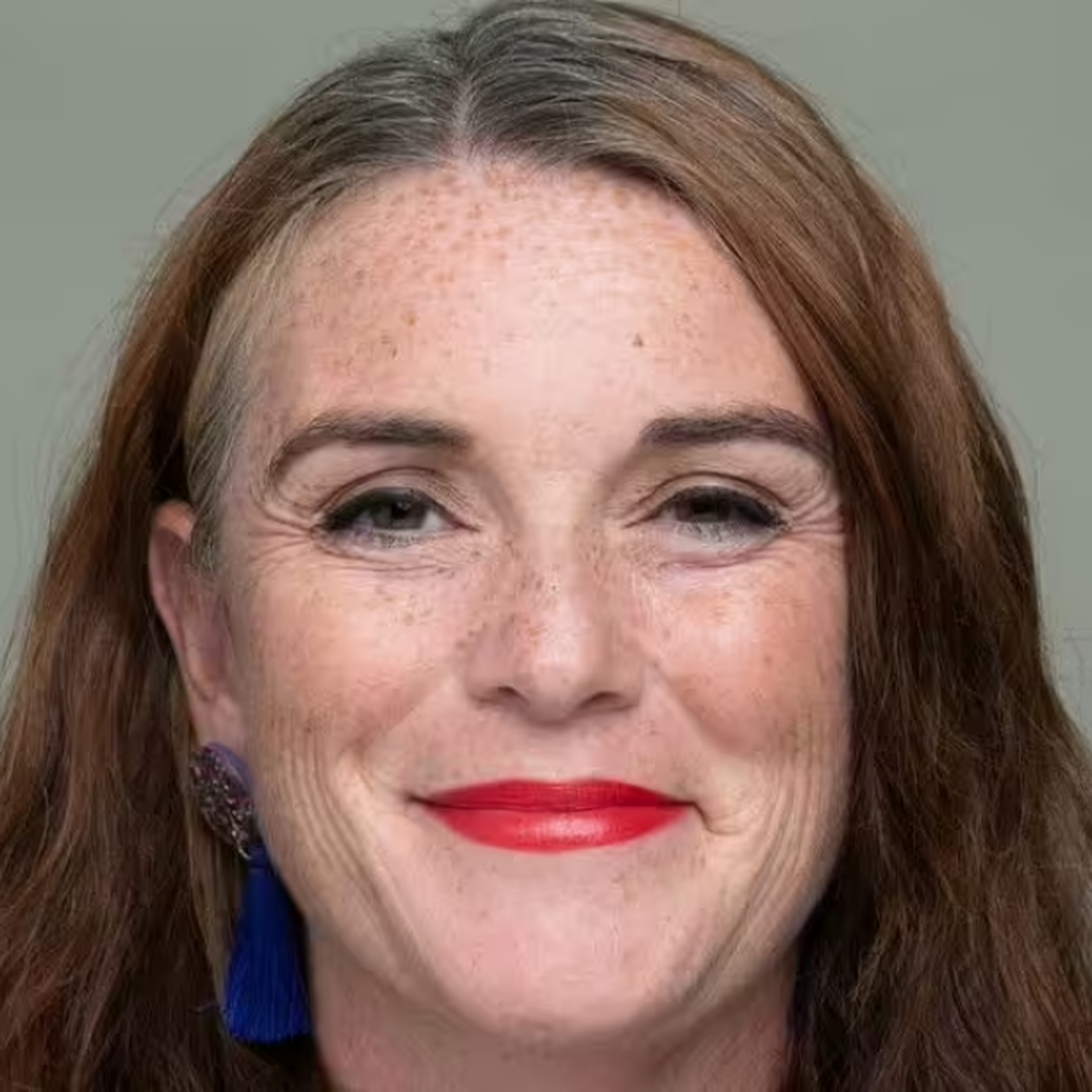 A person with long brown hair and red lipstick smiles at the camera, against a plain background.