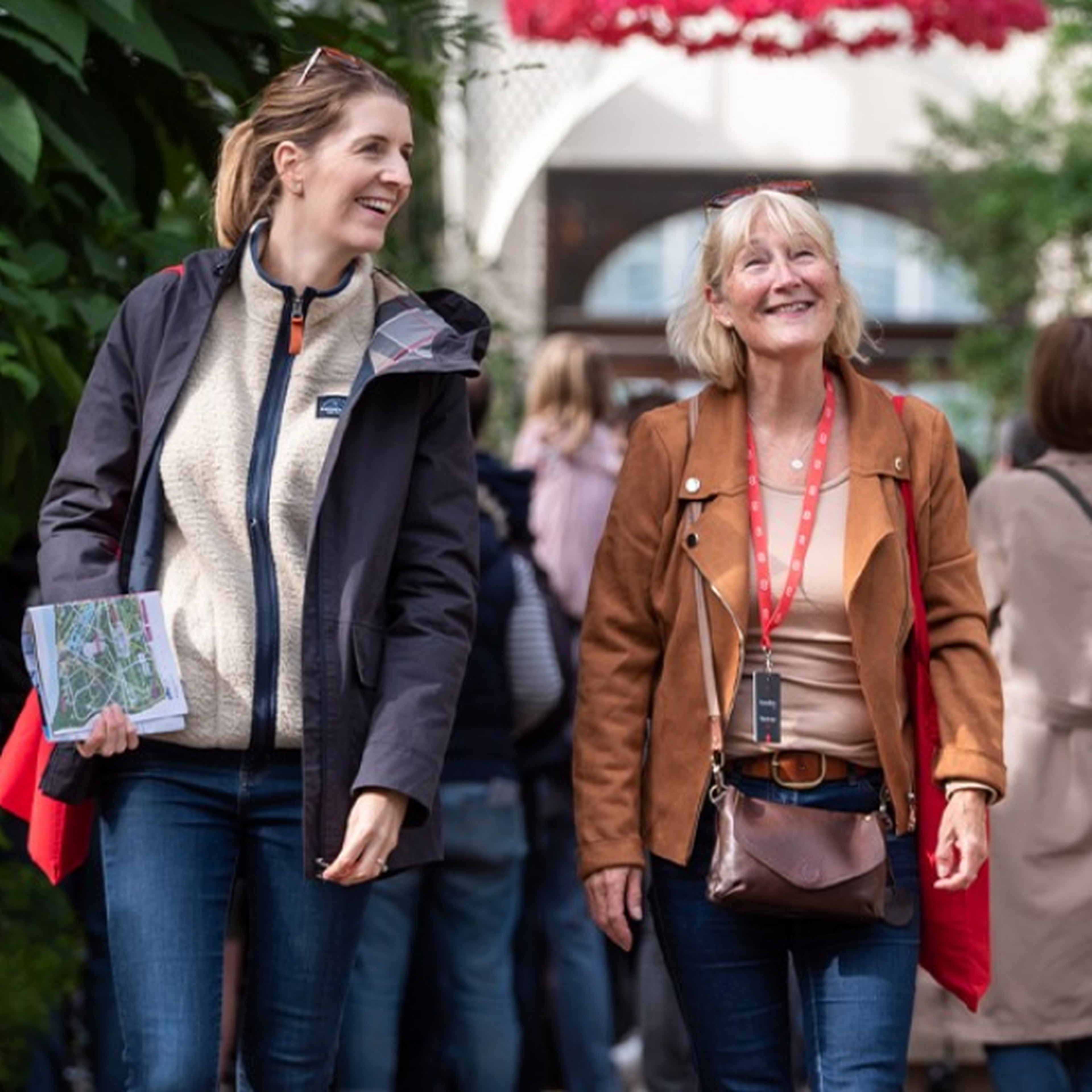 Two women smiling and walking in an outdoor garden area, surrounded by greenery and people in the background.