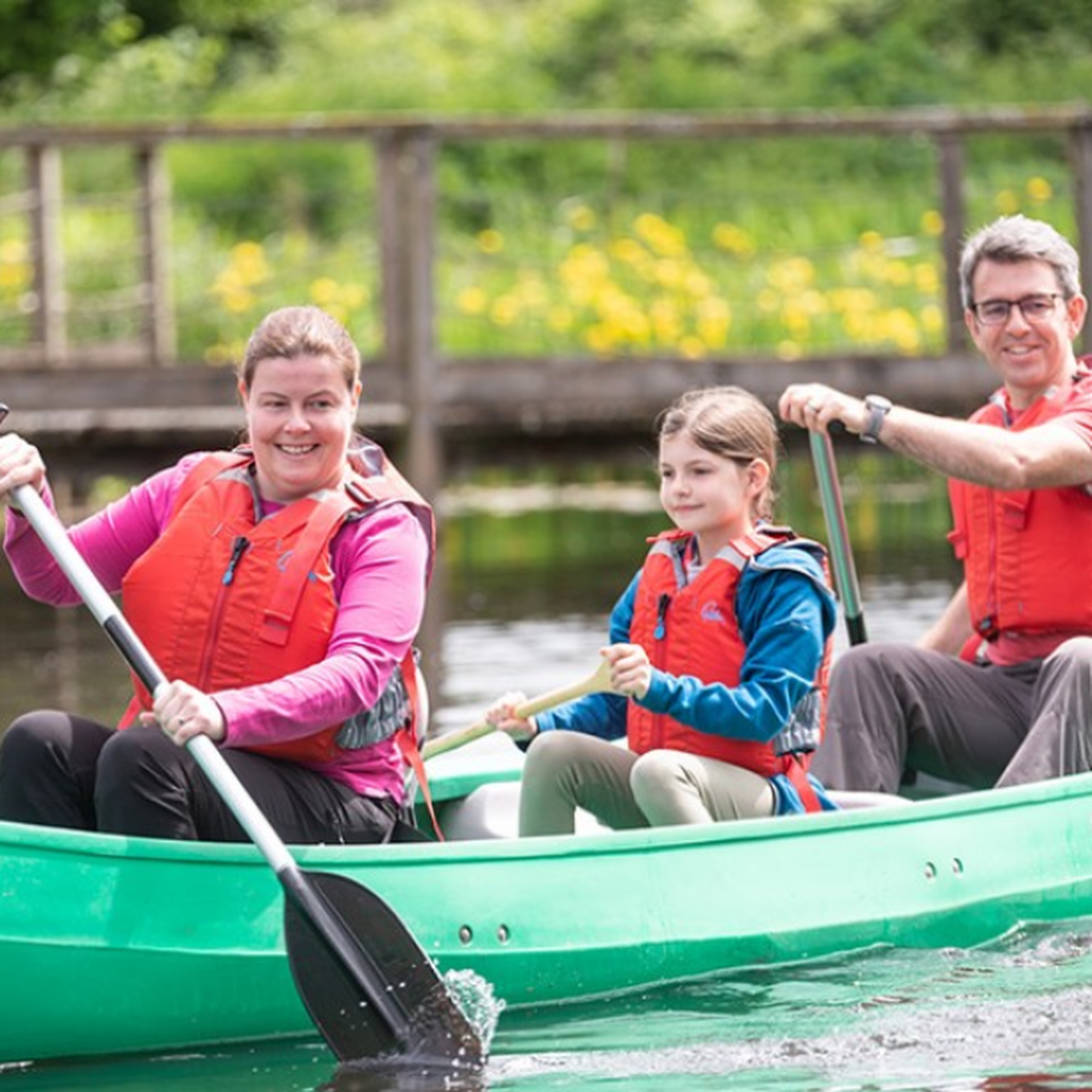 A family of three wearing life jackets paddles a green canoe on a calm river, with a wooden bridge and yellow flowers in the background.