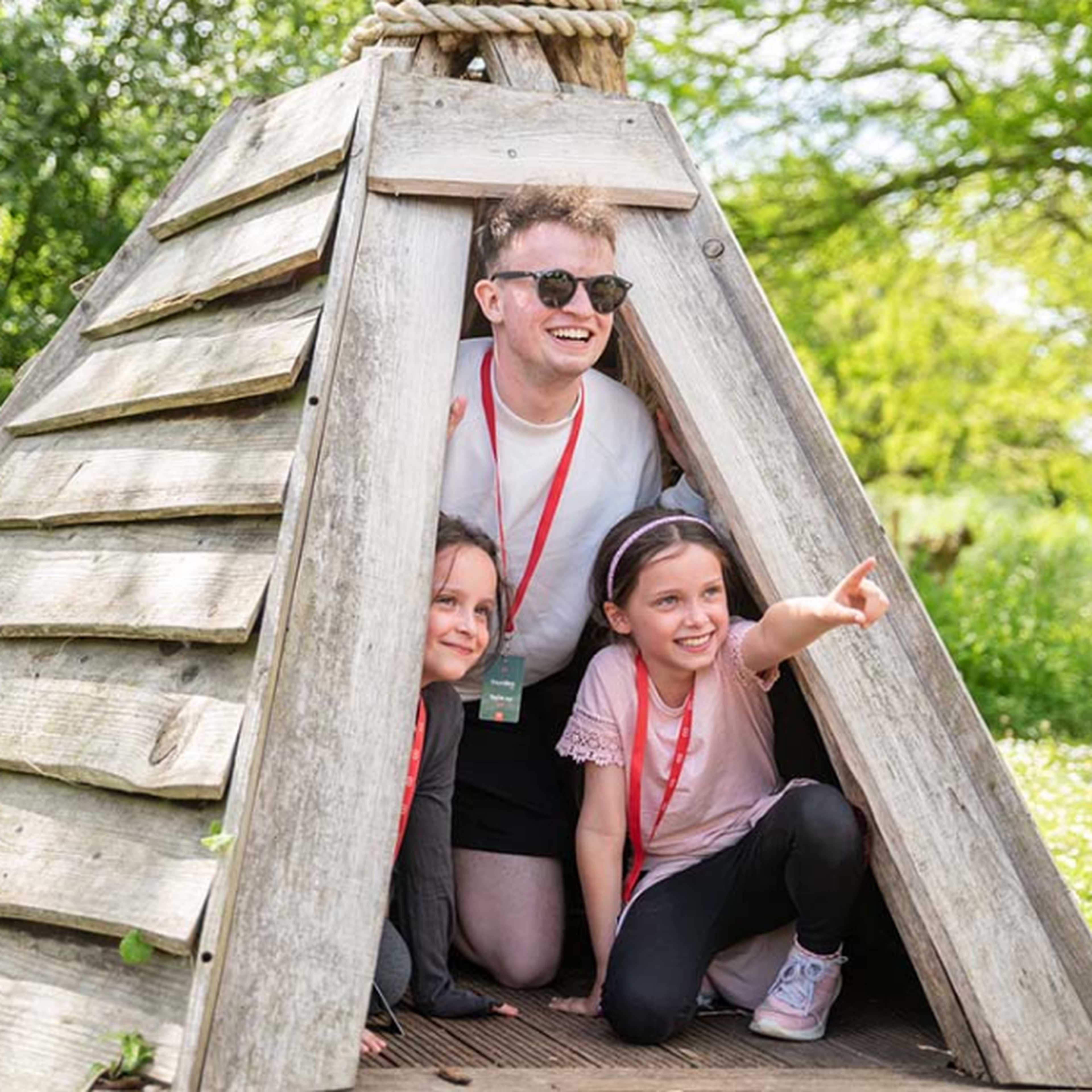 Three people, including two children and an adult, smiling and crouching inside a wooden play structure in a park on a sunny day.
