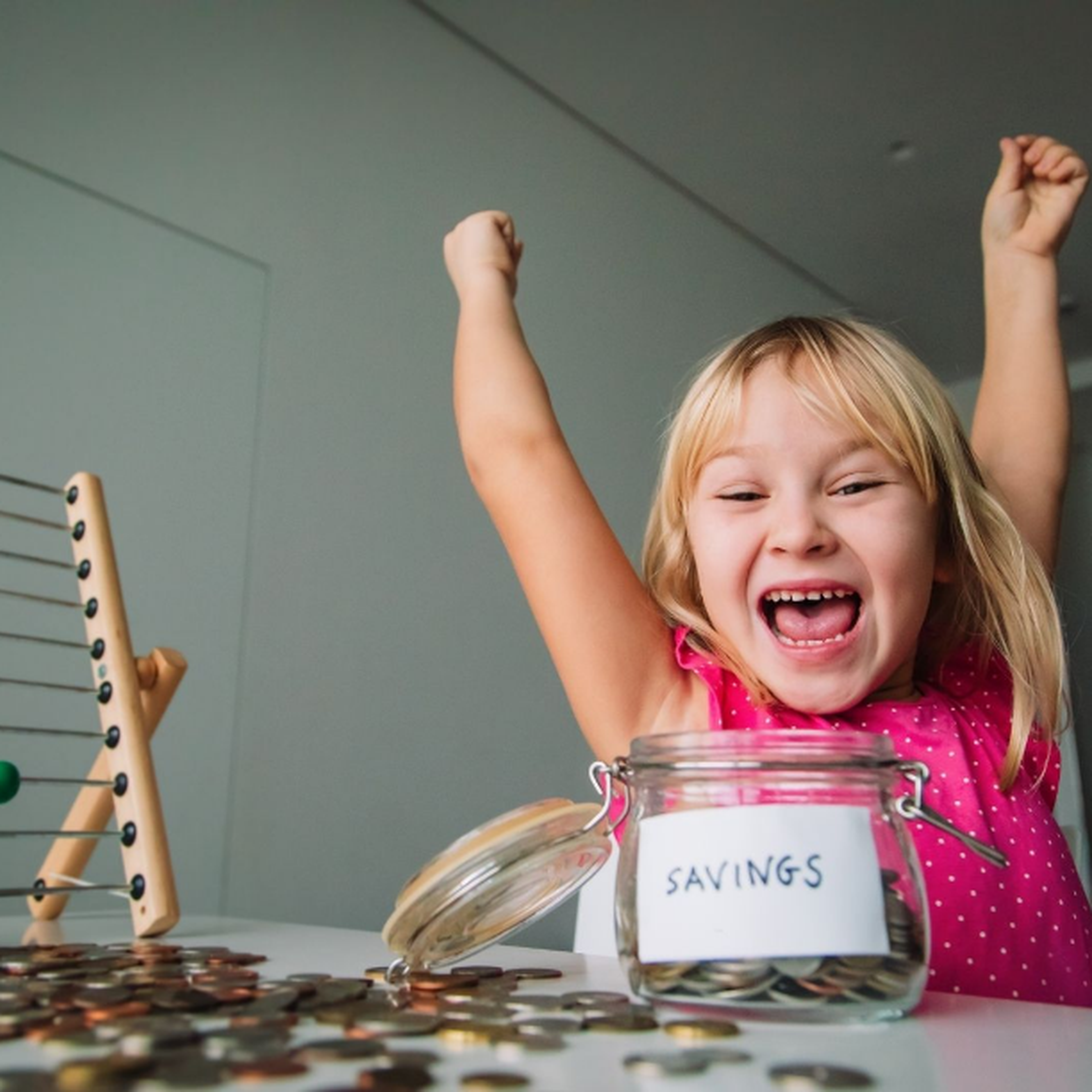 A joyful child with raised arms sits beside a jar labeled "SAVINGS" and a colorful abacus.