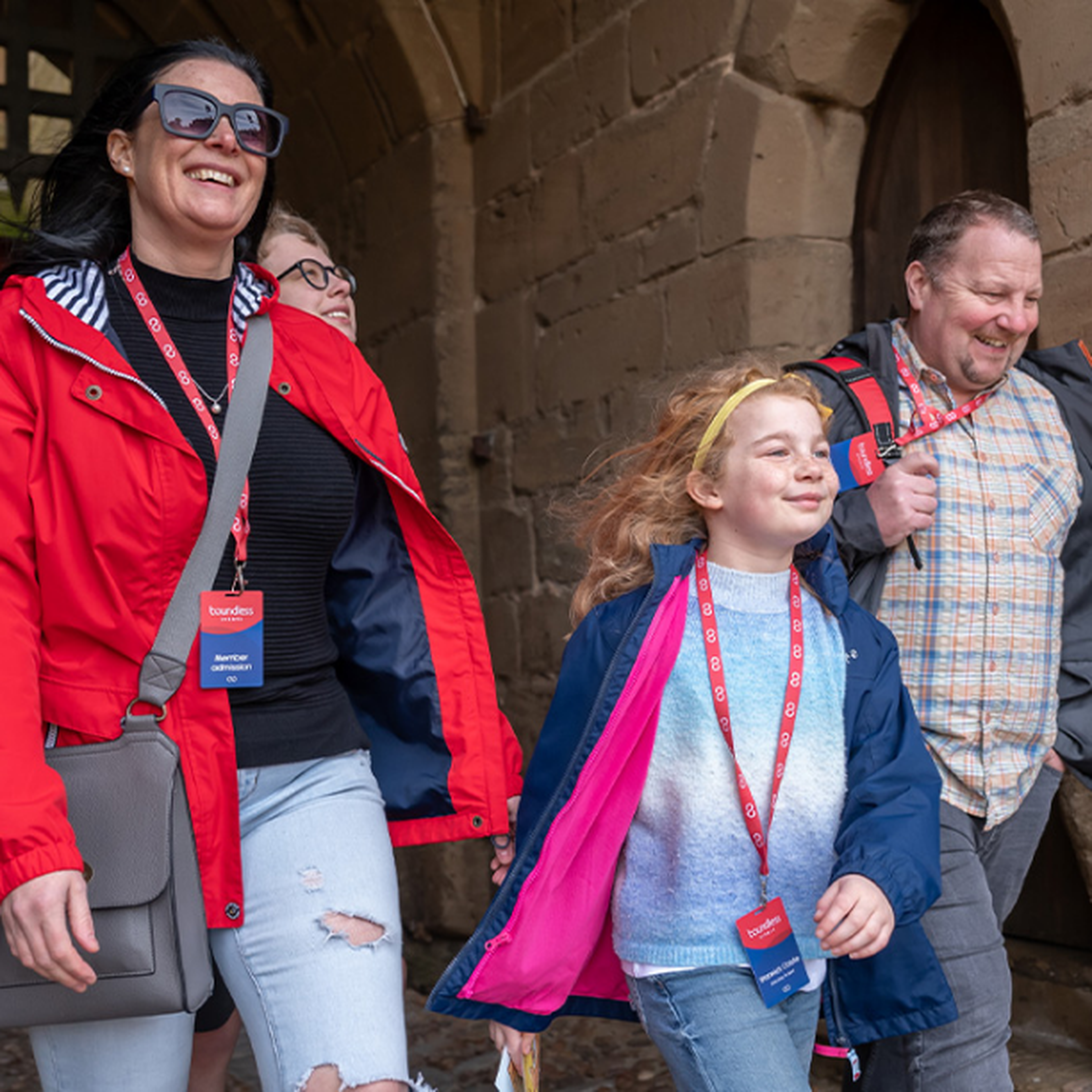 A smiling family in casual clothing walks under an archway, with a woman in a red jacket and a child with a pink cape in front.