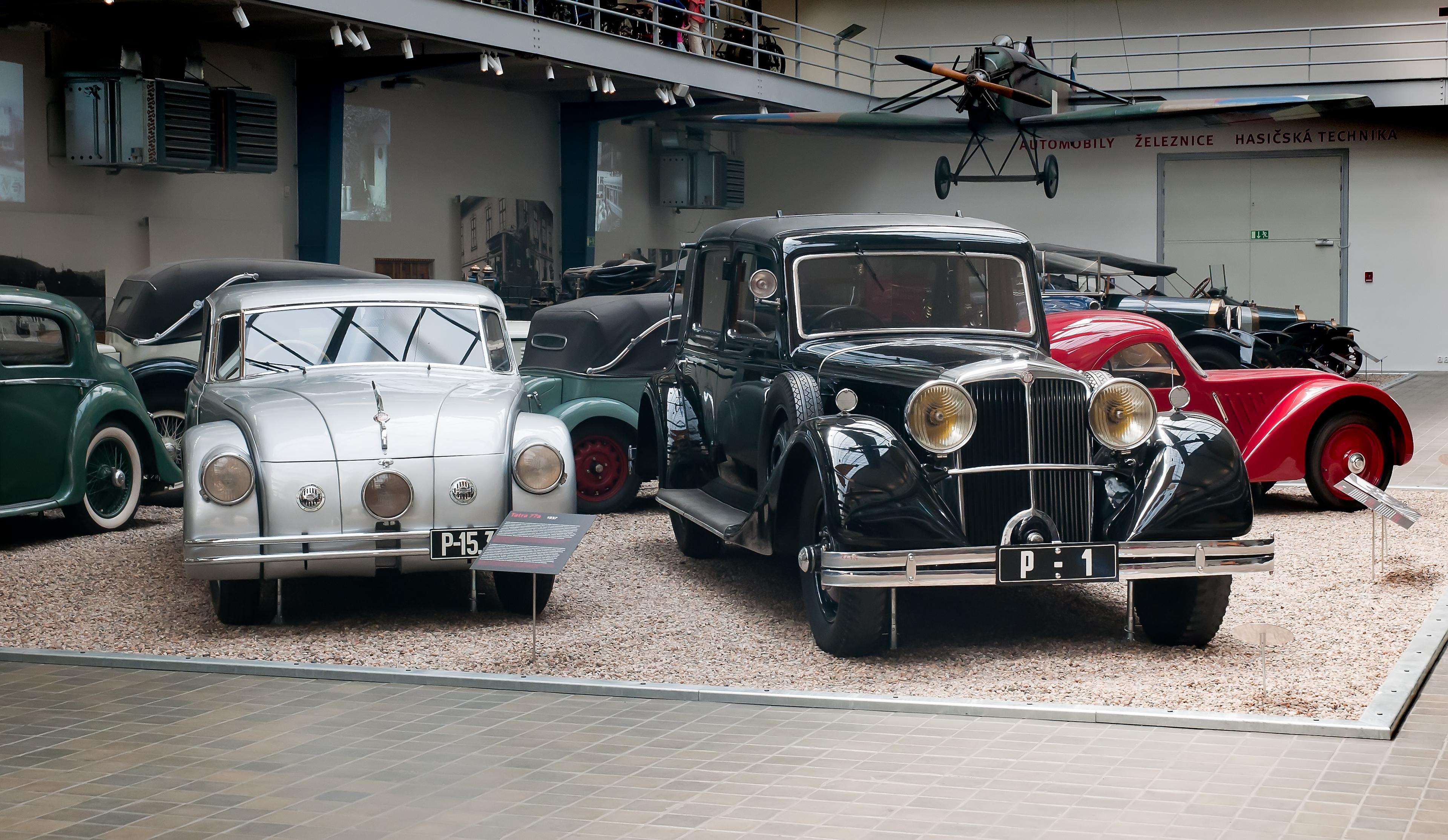 Two vintage cars in a museum display, one silver and one black, with classic designs and a vintage airplane visible in the background.