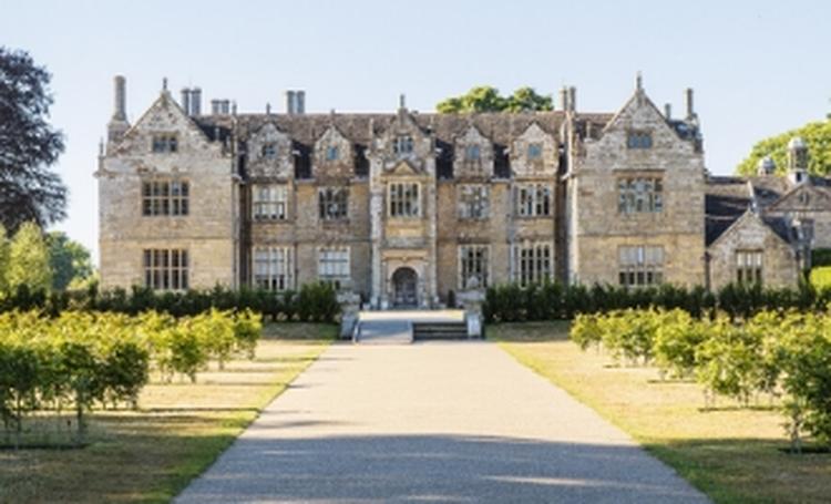 A large, historic stone mansion with multiple gables and tall chimneys, viewed from the end of a path lined with shrubs and greenery.