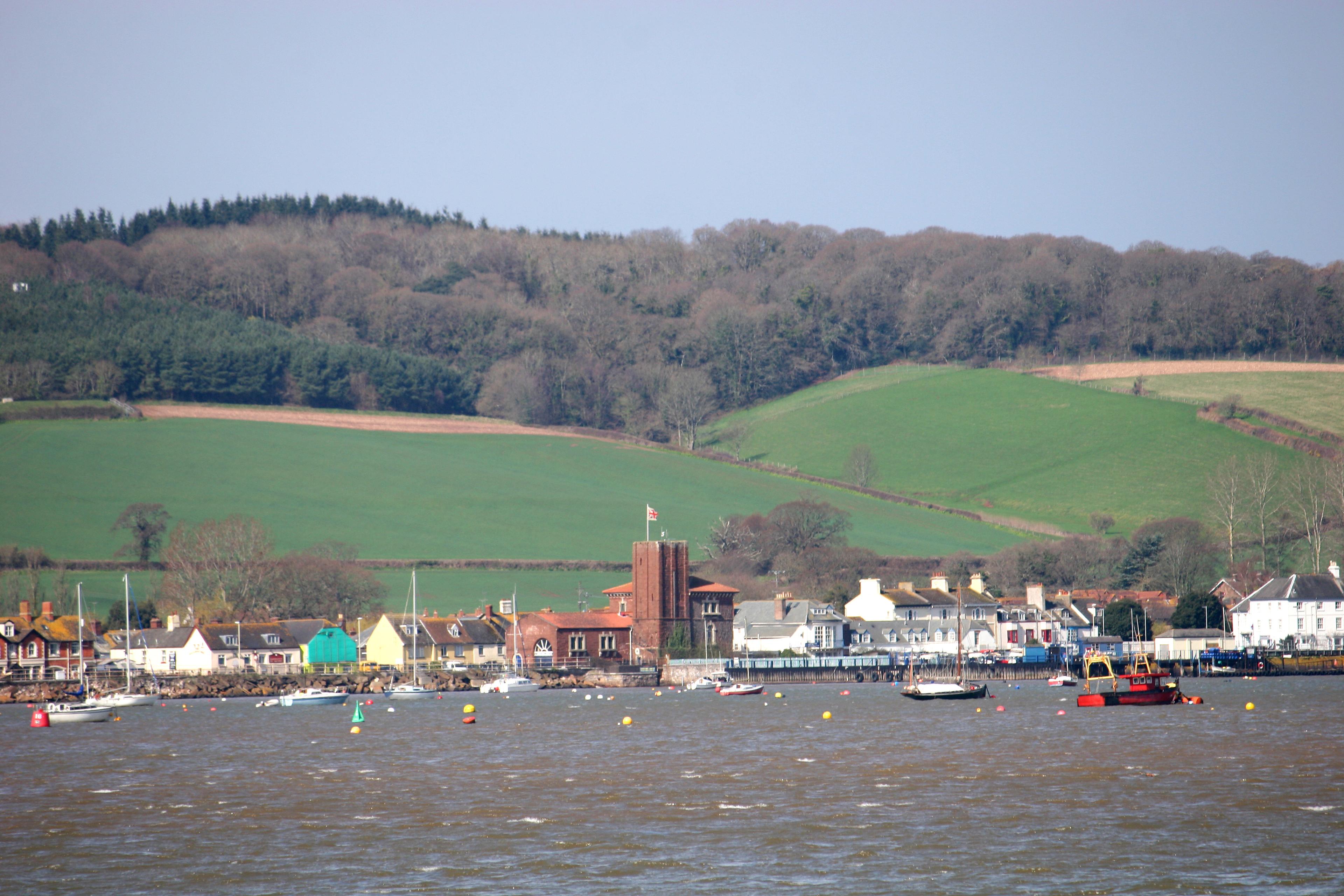 A tranquil coastal village and harbor with colorful boats, a church tower, and green rolling hills beneath a pale blue sky.