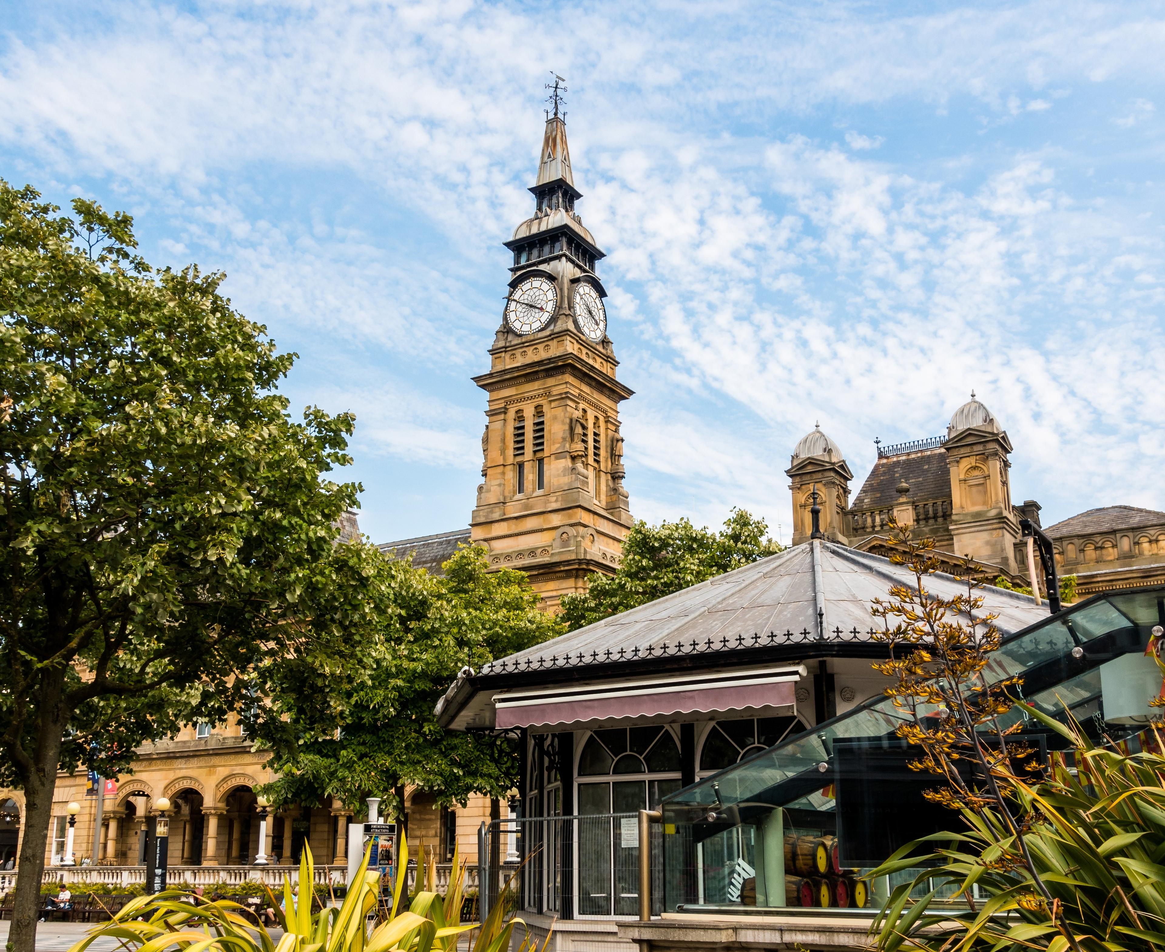 Historic clock tower with a pointed roof and clock face, surrounded by trees and modern structures, under a partly cloudy sky.