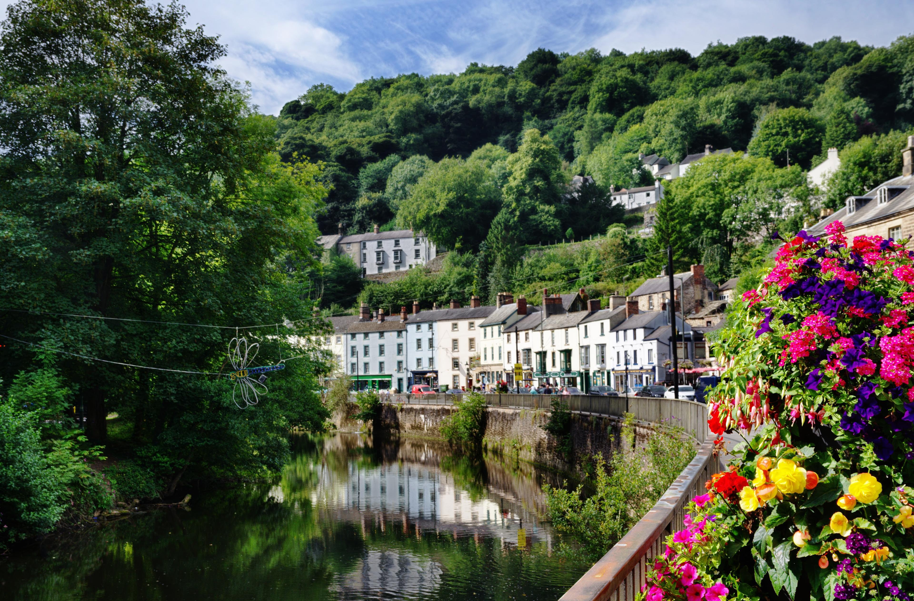 Quaint village by a river with colorful hillside buildings, lush trees, and vibrant flowers under a blue sky.