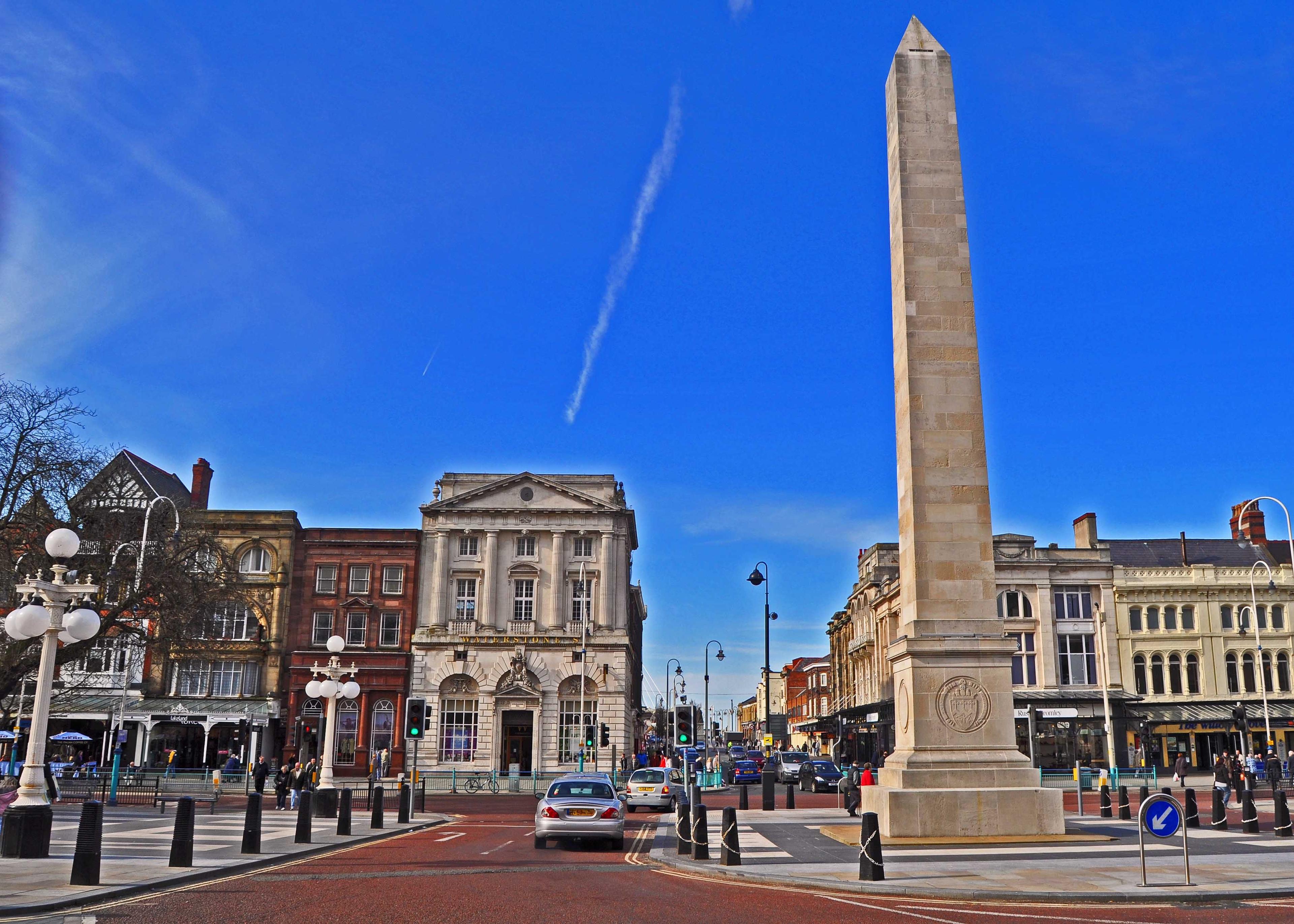 Urban square with a tall obelisk, historic buildings, cars, and people. A clear blue sky and trees frame the scene.