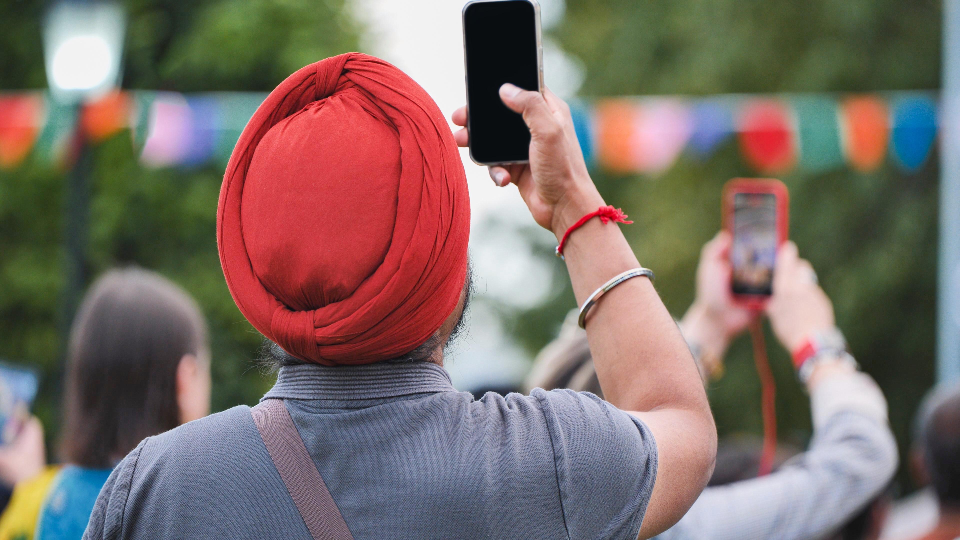 A person wearing a red turban holds up a smartphone, capturing a scene at an outdoor event with colorful flags in the background.