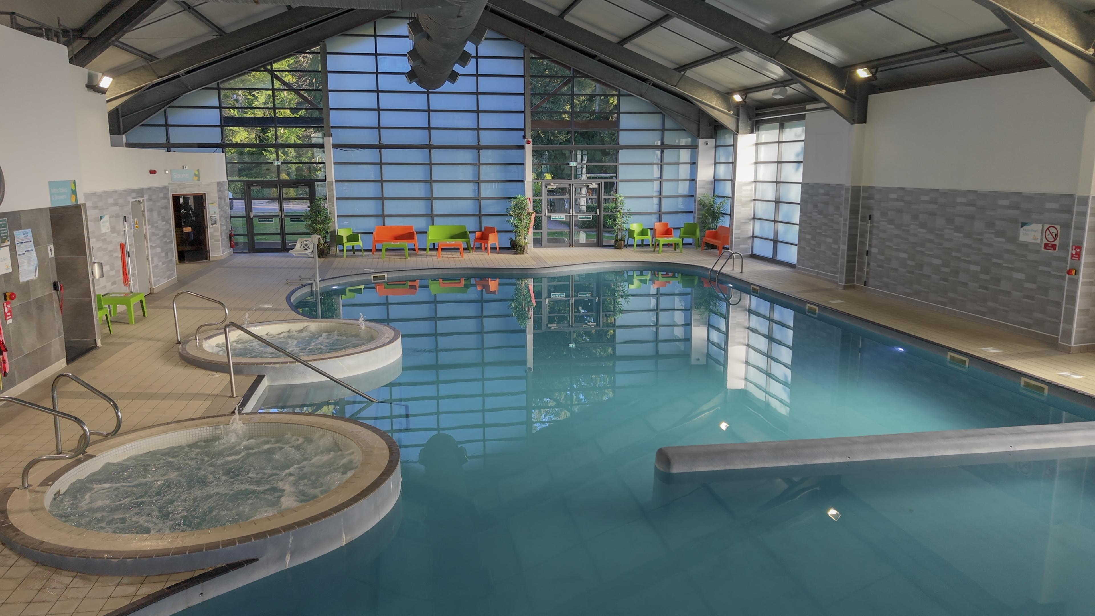 Indoor swimming pool with large windows, two round hot tubs, and colorful chairs lining the deck.
