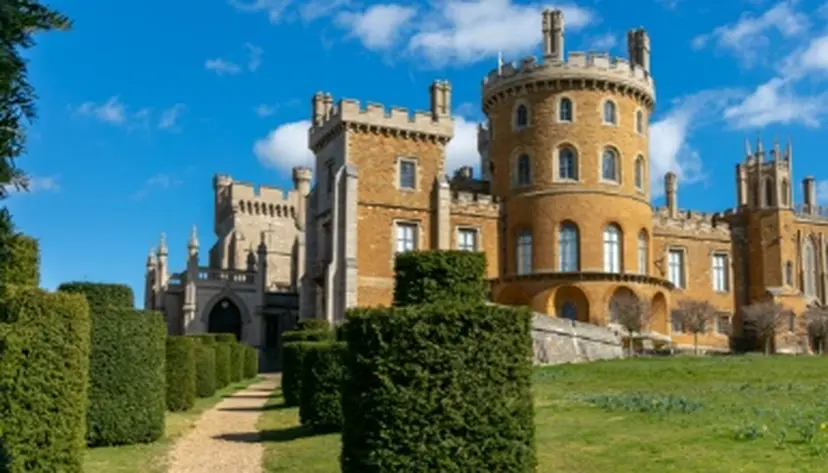 Historic castle with round towers and battlements, set against a blue sky. Manicured hedges and a gravel path lead to the entrance.