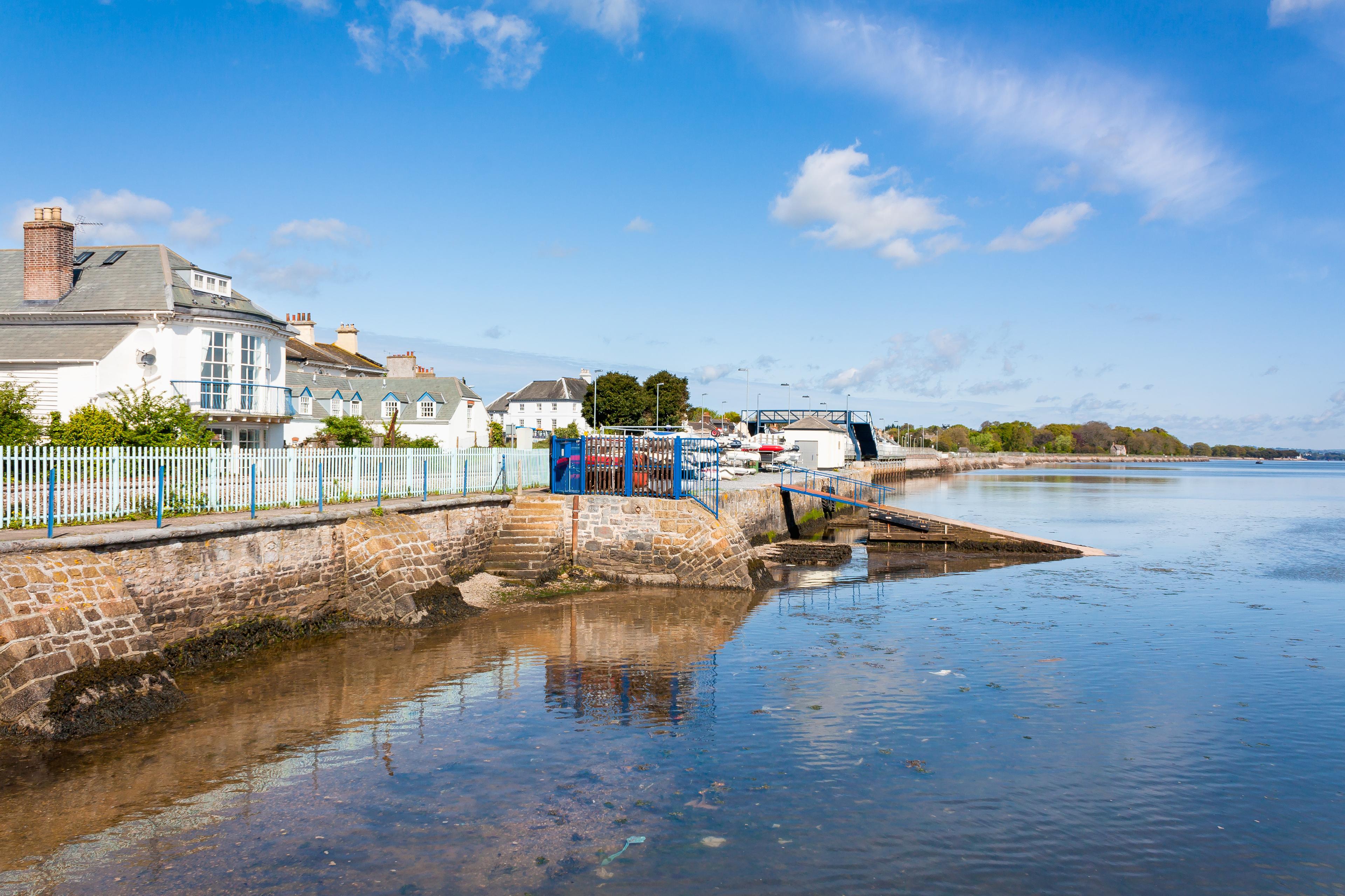 Bright coastal harbor with white cottages, stone seawall, colorful boats, and calm blue water beneath a sunny, cloud-dotted sky.