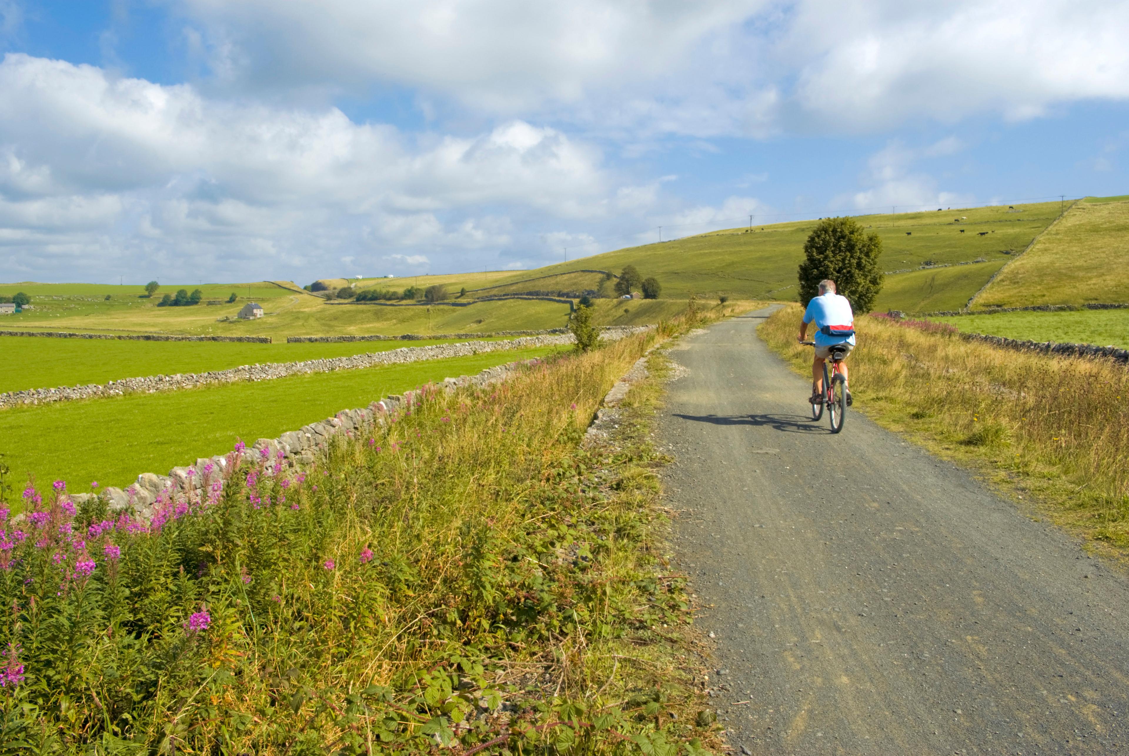 A cyclist rides along a rural dirt path, surrounded by green fields and stone walls under a partly cloudy sky.