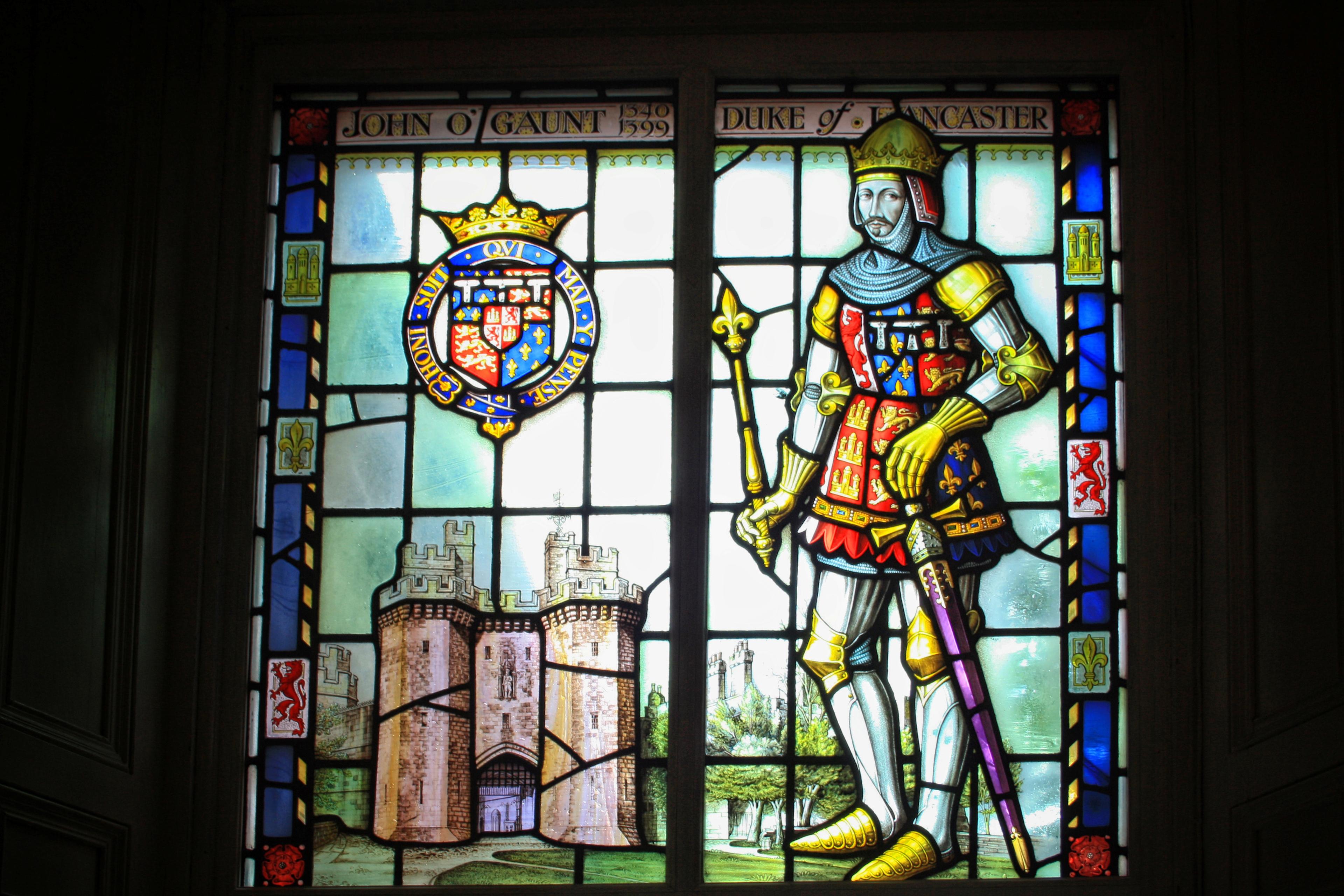 Stained glass depicting John of Gaunt in armor, with a coat of arms and a castle scene, bordered by intricate designs.