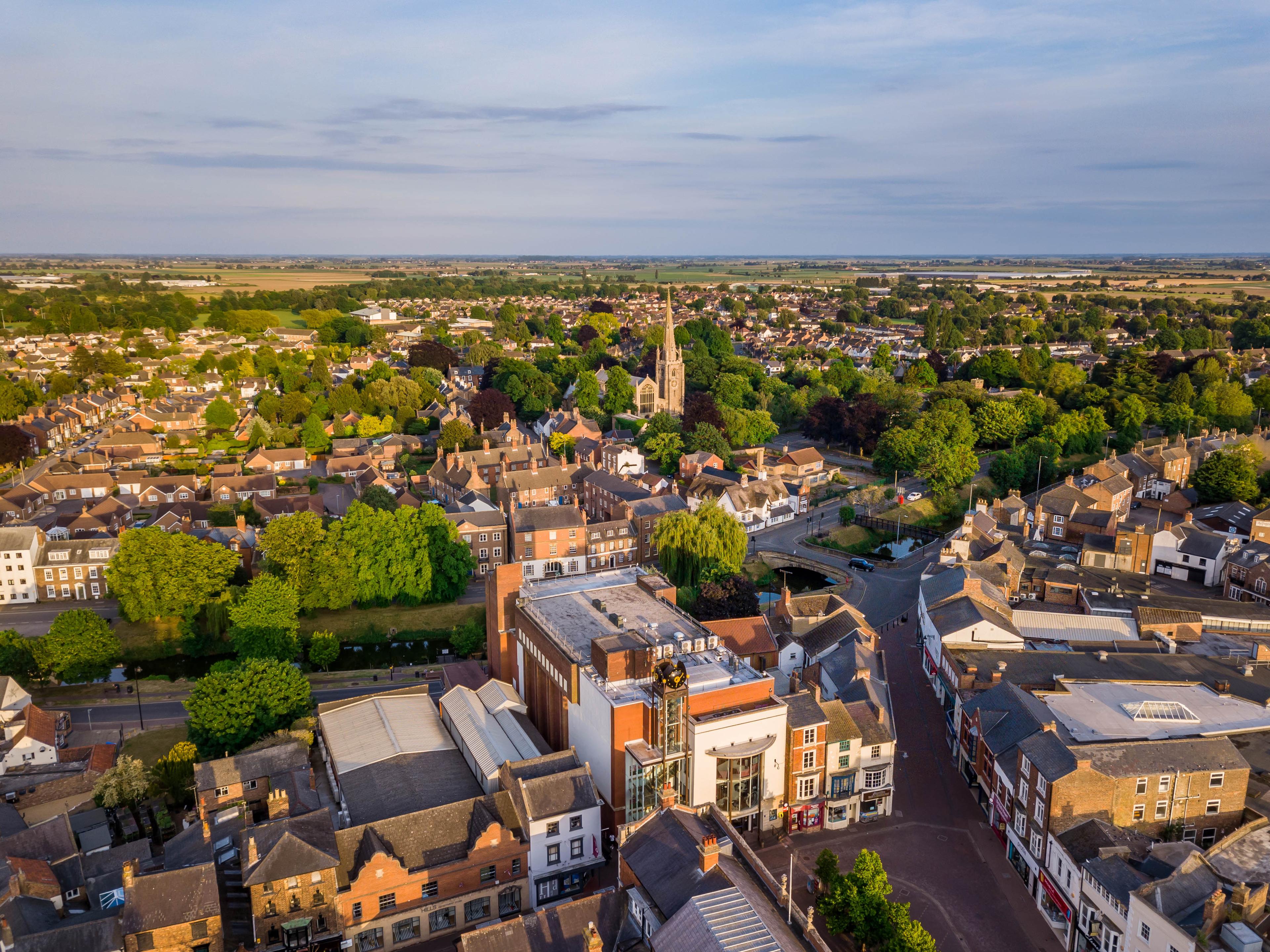 Aerial view of a quaint town with a mix of historic and modern buildings, a church spire, lush greenery, and winding streets under a clear sky.