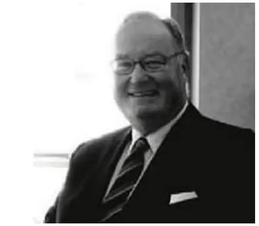 Smiling man in a suit and tie, wearing glasses, stands beside a window in a black and white photo.
