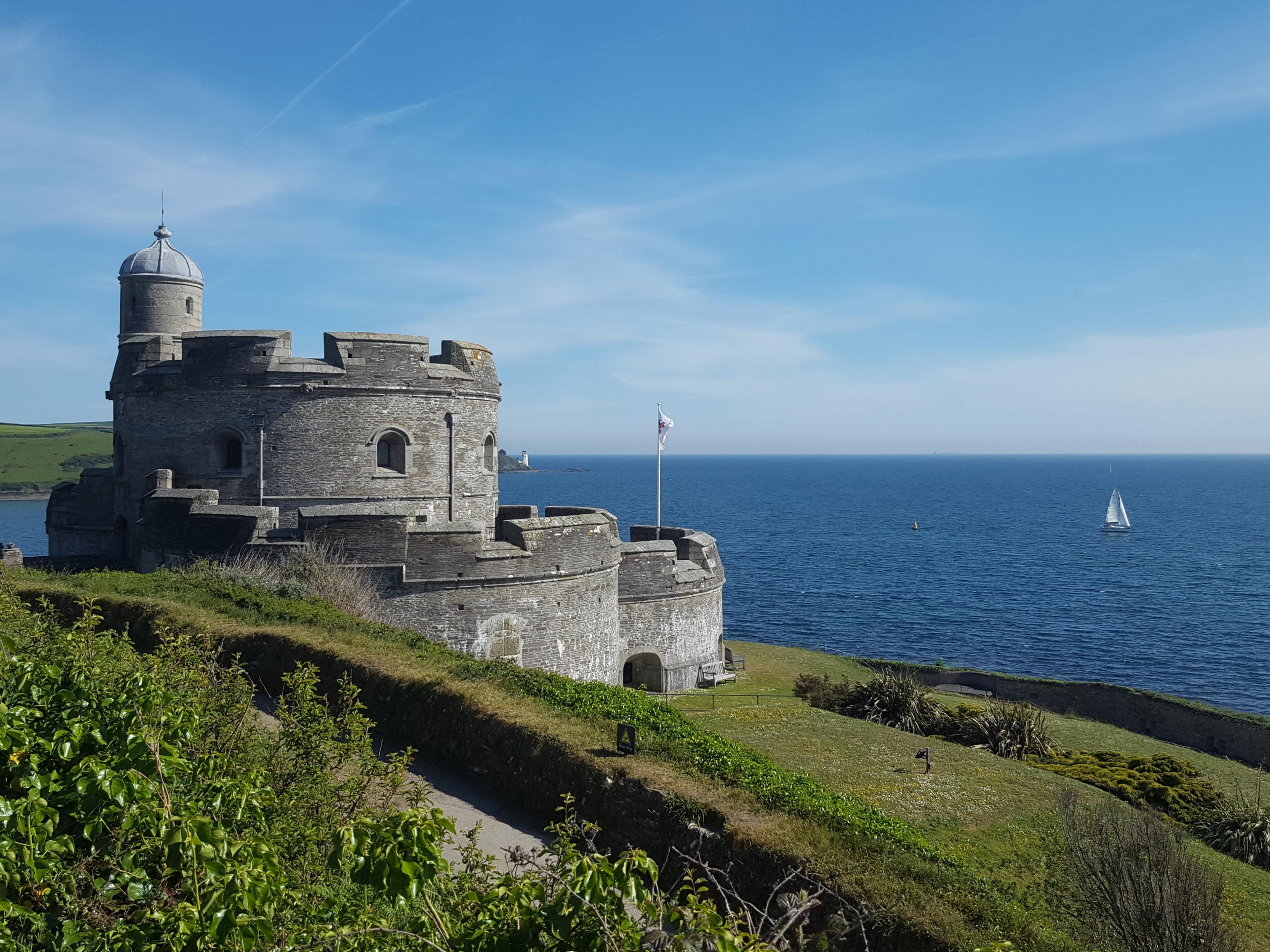 A historic stone castle overlooks the blue sea under a clear sky, with a sailboat in the distance. Greenery surrounds the foreground.