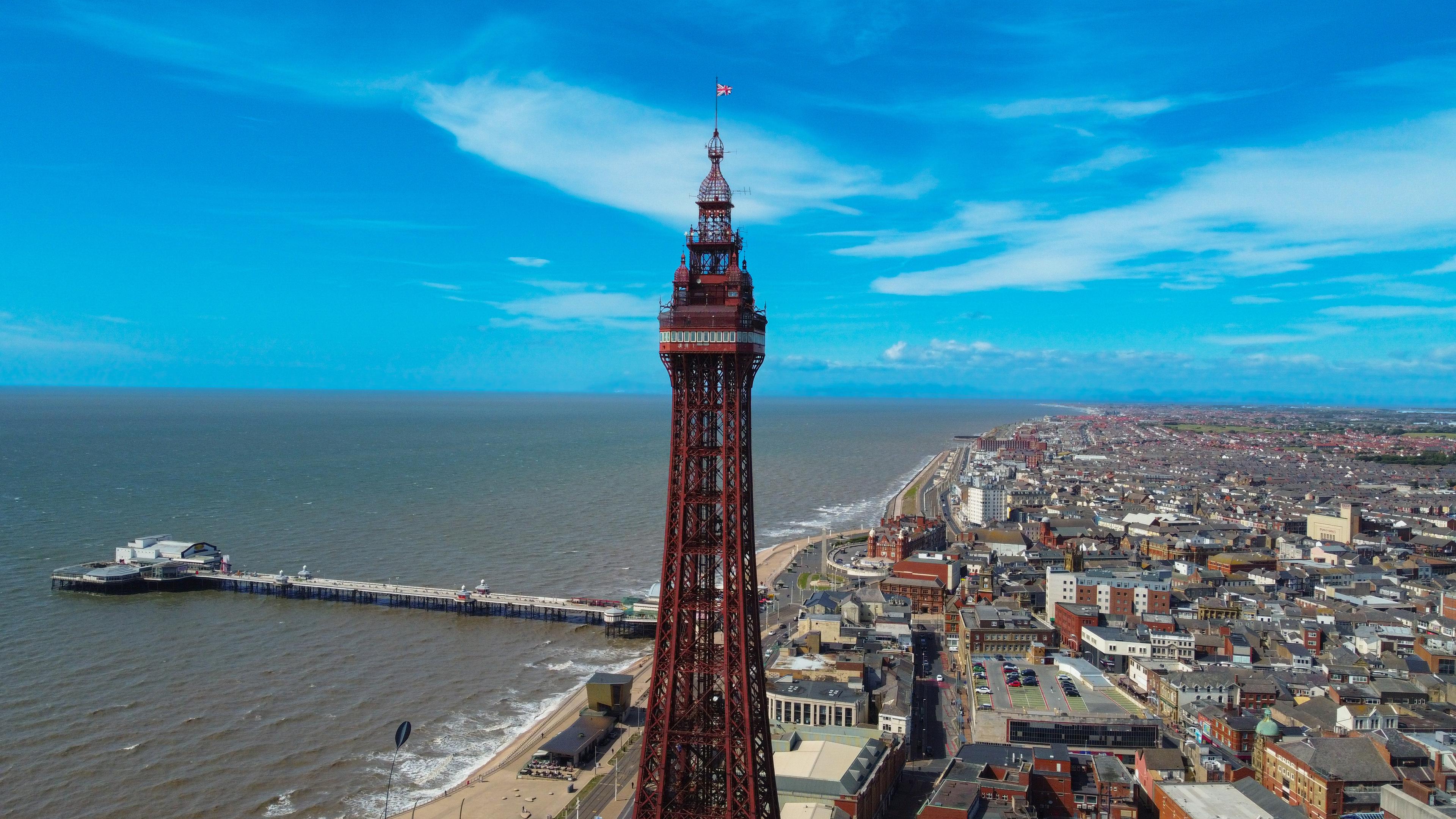 A tall, red tower stands beside a beach, overlooking a coastal city and pier under a bright blue sky with scattered clouds.