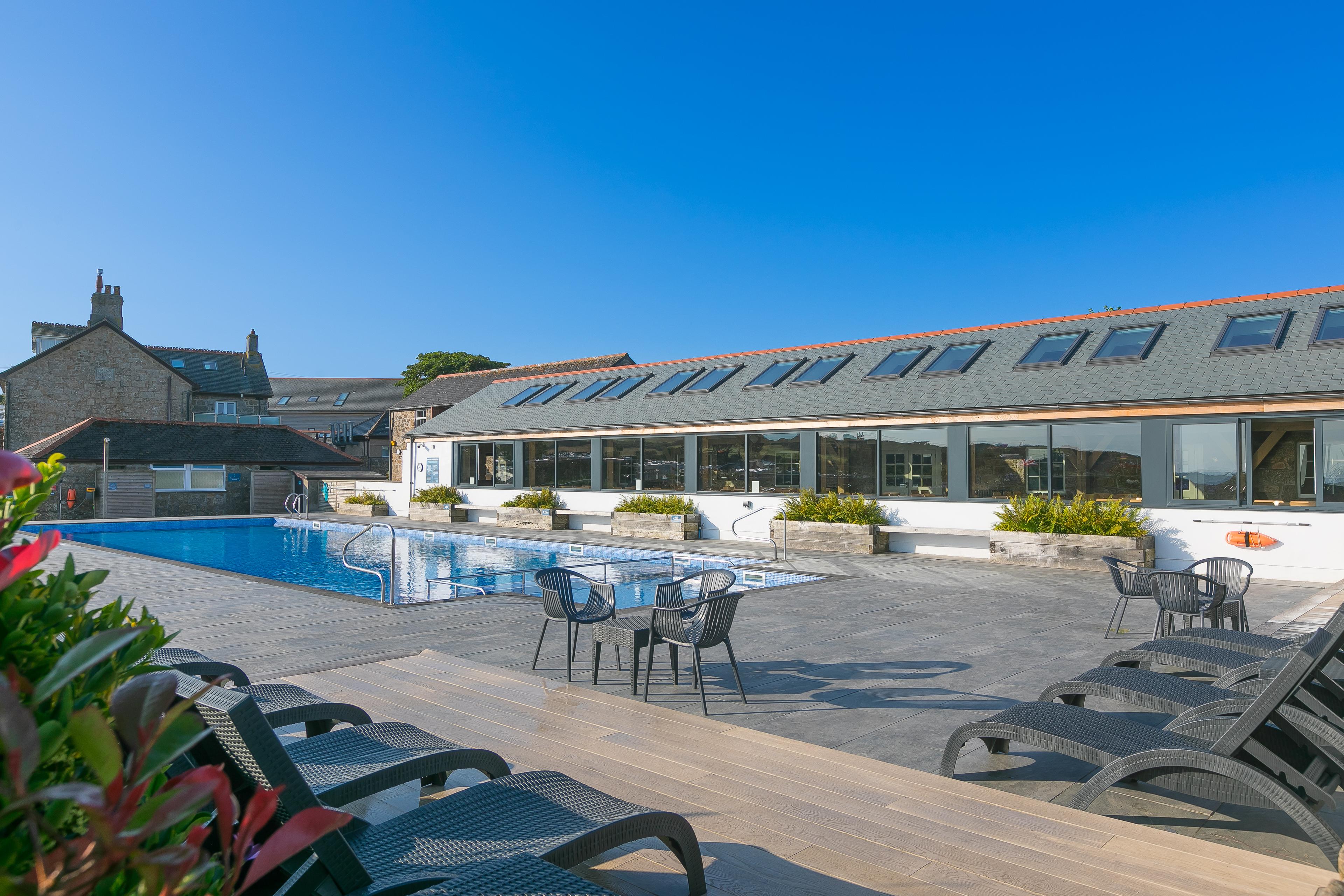 Outdoor pool with sun loungers and chairs, surrounded by a modern building with skylights under a clear blue sky.